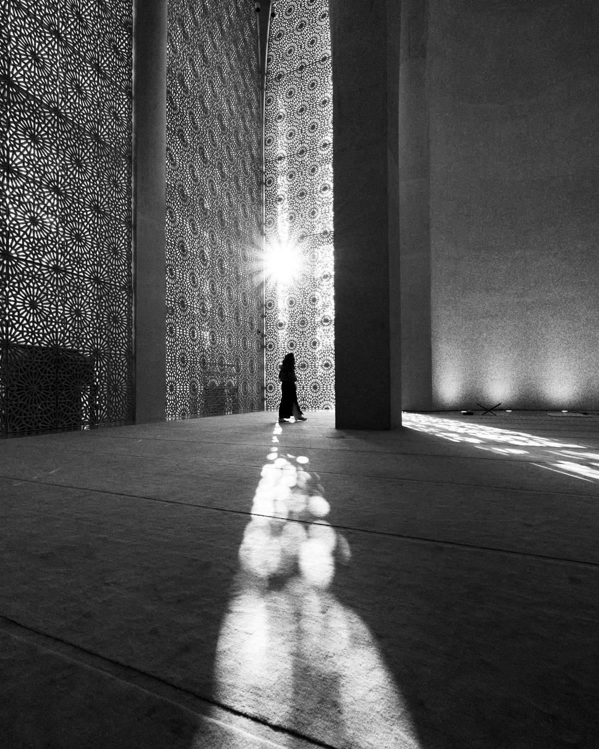 A person walking inside a modern building with large decorative lattice panels, sunlight streaming through, creating shadows on the floor.