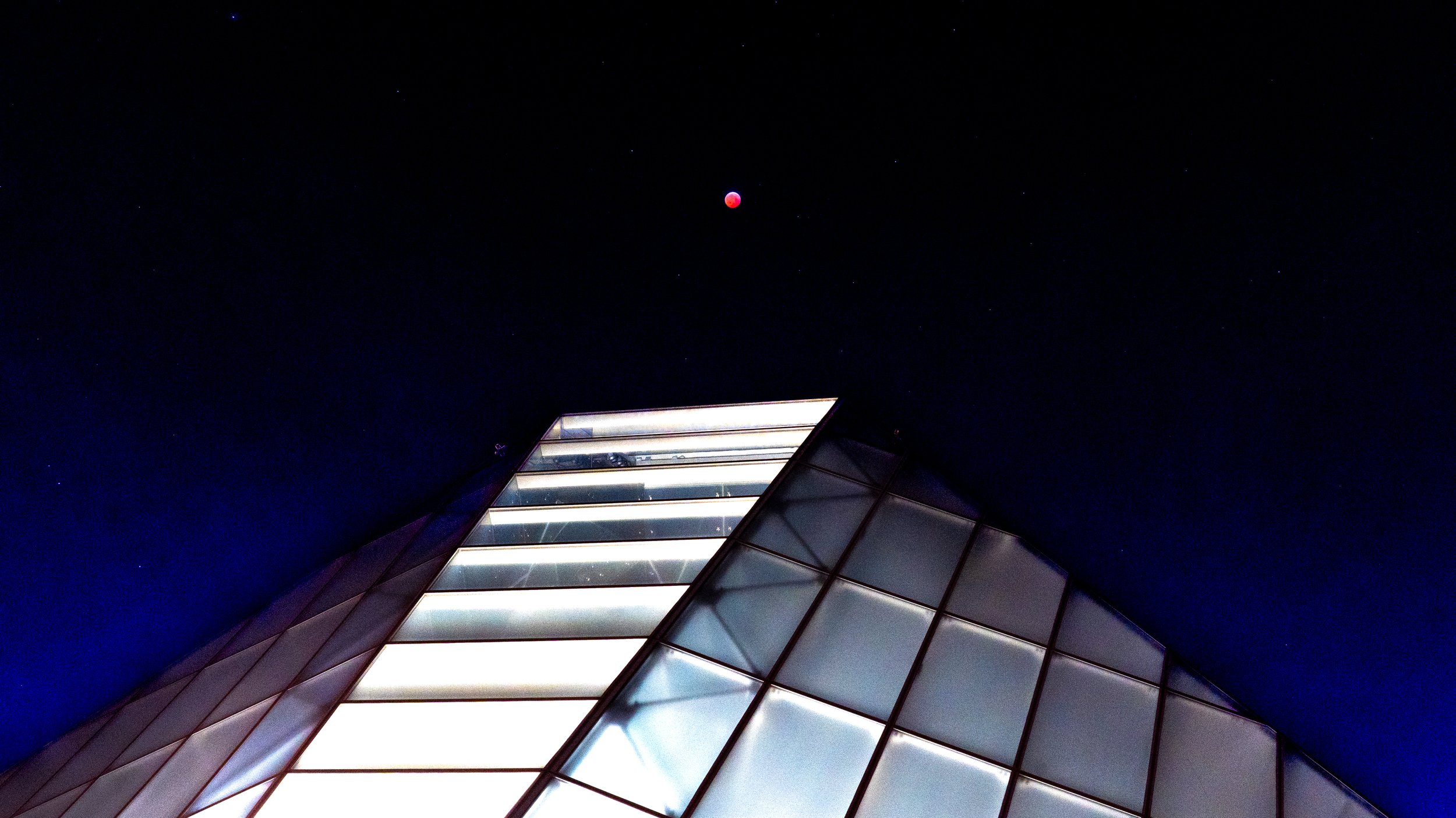 Nighttime view of a modern glass building with illuminated panels and a clear, dark sky with stars and a red moon above.