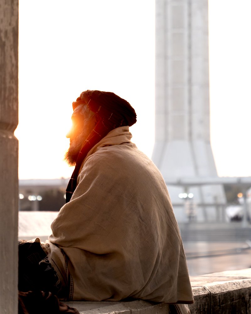 A man with a beard sitting at a window seat, with sunlight shining on his face, in front of a rocket or launch pad.