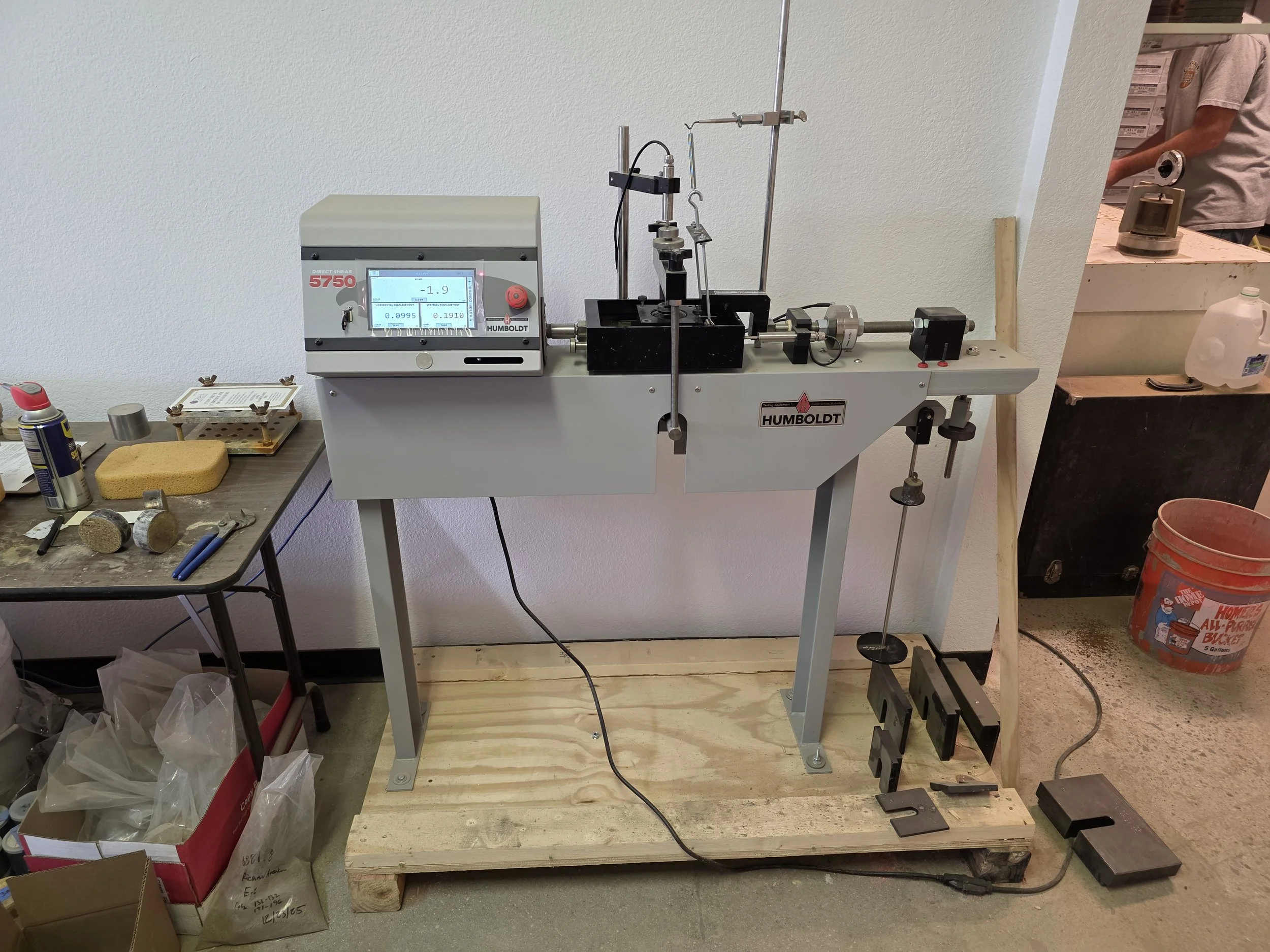A laboratory apparatus setup with a Humboldt direct shear testing device on a wooden frame, connected to various metal rods and clamps. To the left, a workbench has tools, a sponge, and miscellaneous items.