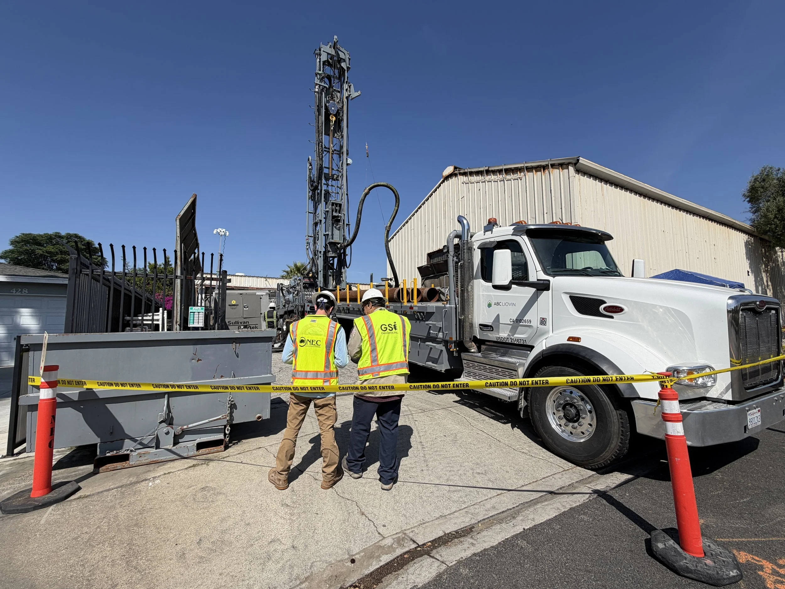 Workers and engineers in safety vests and helmets inspecting a drilling or construction vehicle behind a caution tape barrier at a construction site, with a building and a clear blue sky in the background.