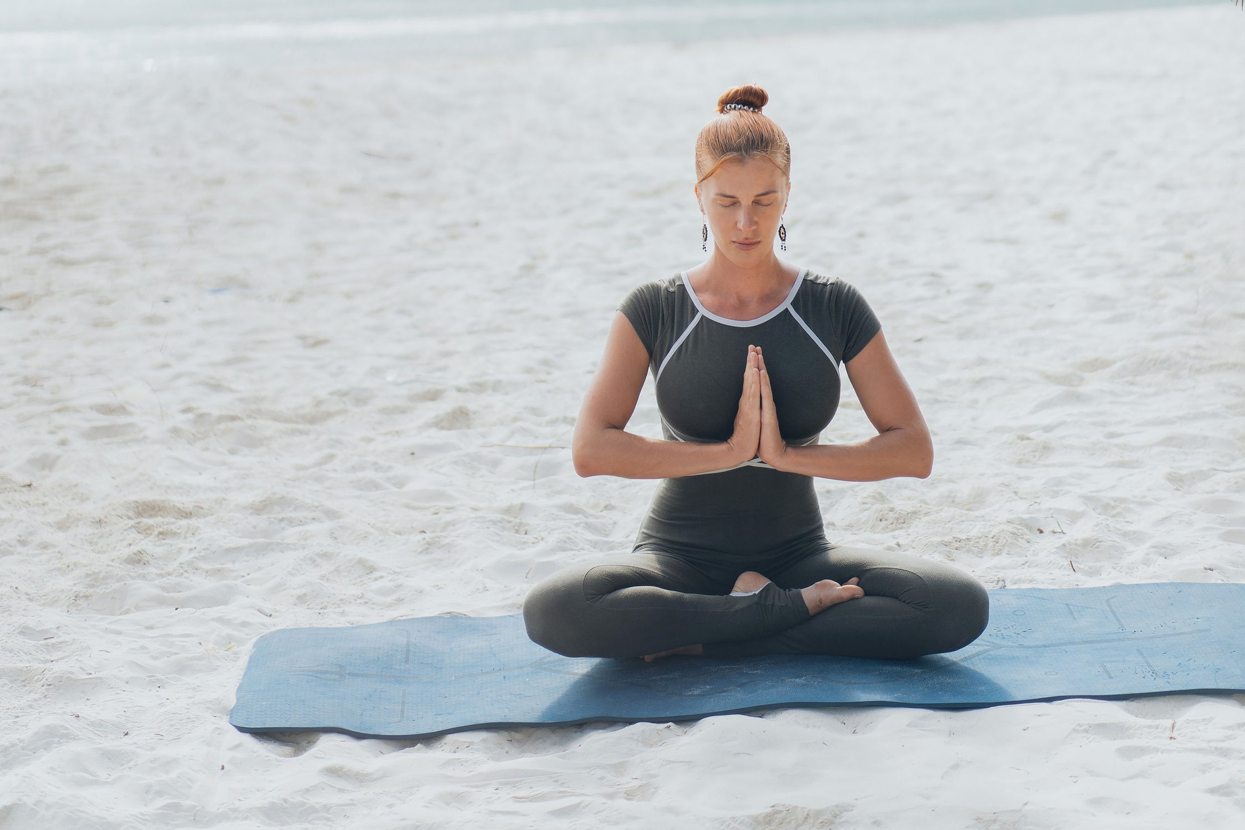 Focused woman in her forties meditating in a cross-legged position on a yoga mat at the beach — cultivating inner peace, body awareness, and mind-body alignment.