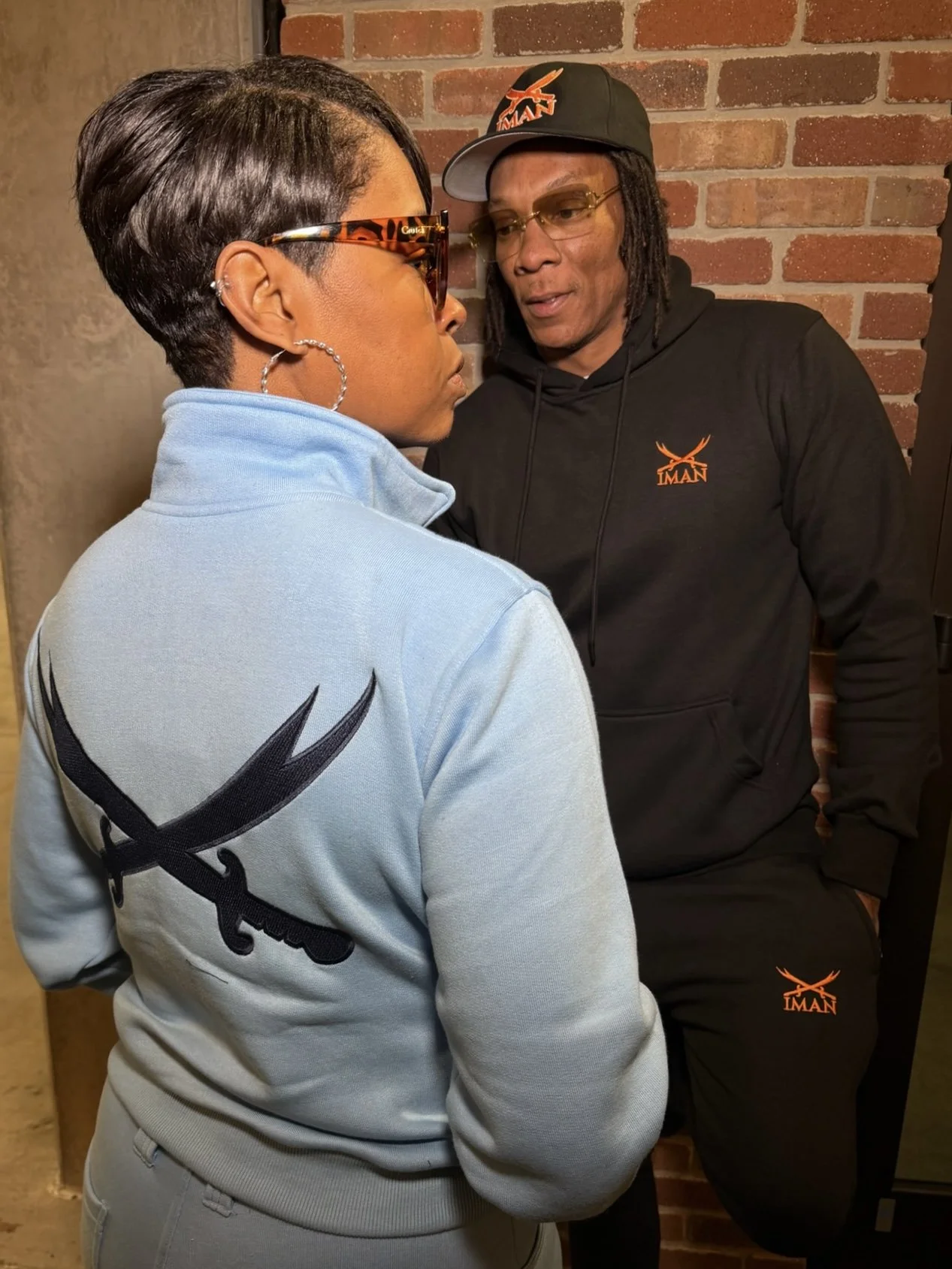 Two people facing each other, a woman with short dark hair wearing sunglasses, hoop earrings, and a light blue jacket with a black embroidered eagle logo, and a man with dreadlocks wearing glasses, a black hoodie, and a black cap with an orange logo,