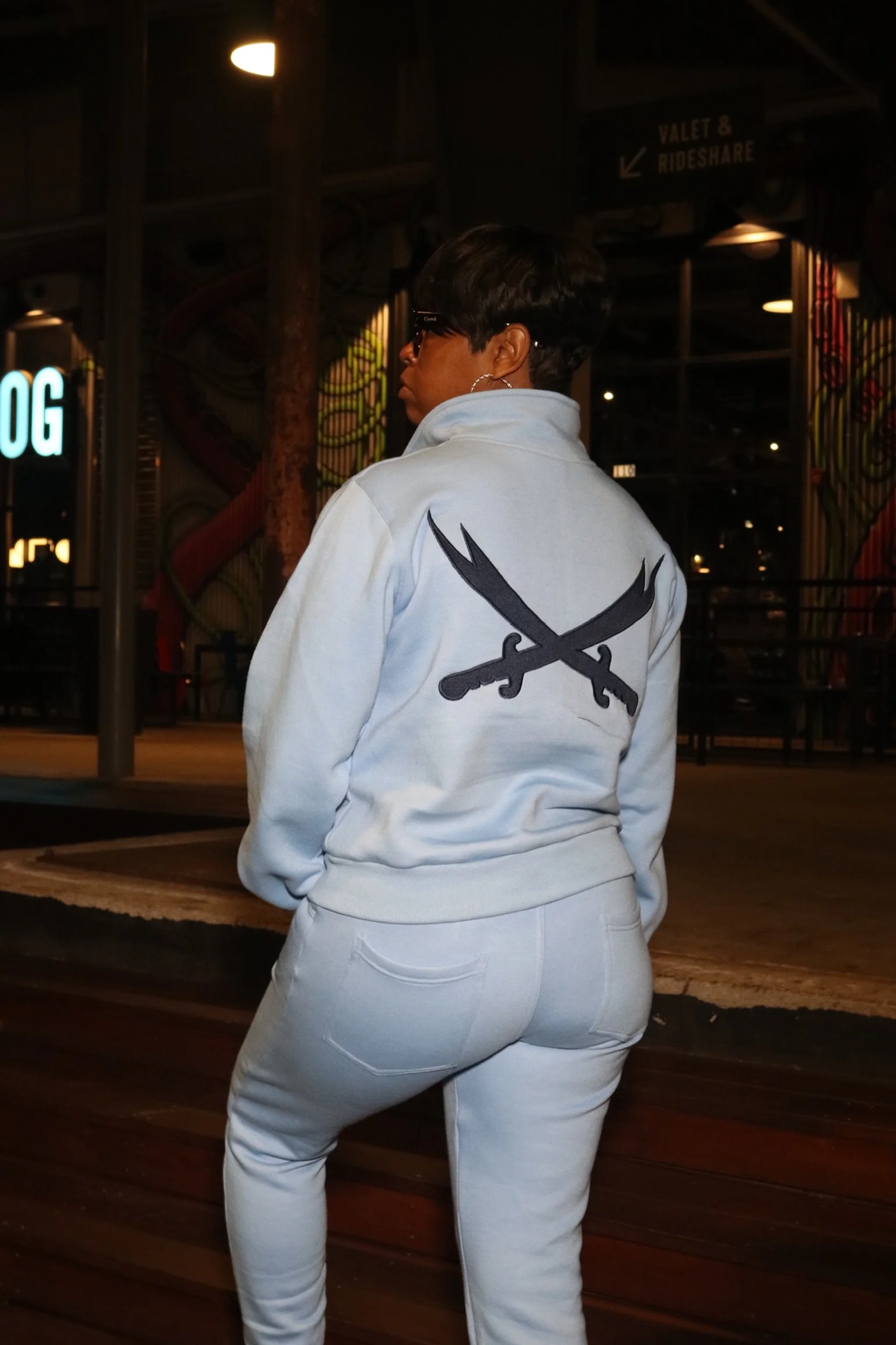 Person in a white jacket with crossed black swords emblem on the back, standing outside at night.