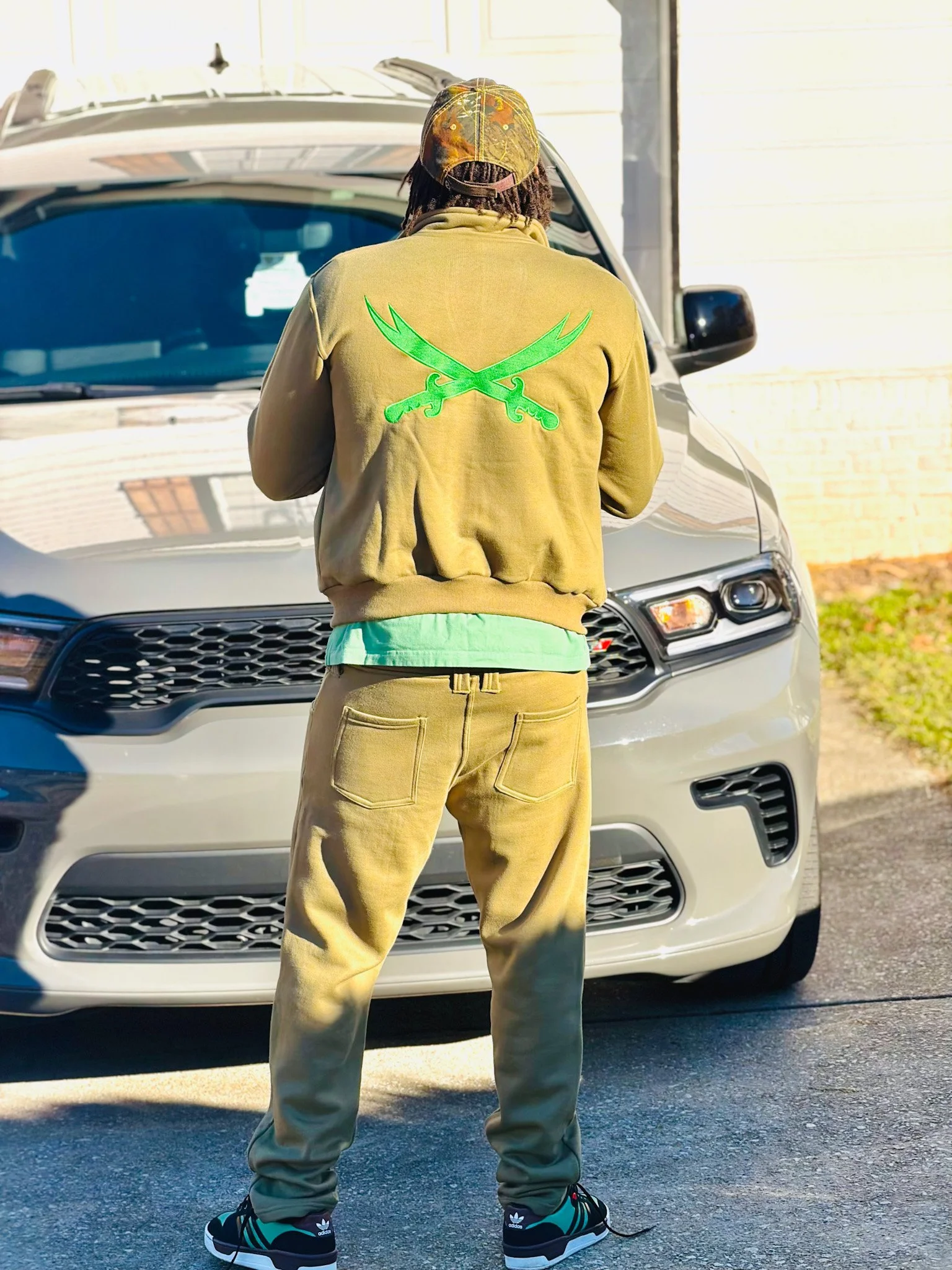 Person standing in front of a gray car, seen from behind, wearing a tan hoodie with green crossed swords on the back, tan pants, a colorful cap, and black sneakers with teal and white accents.