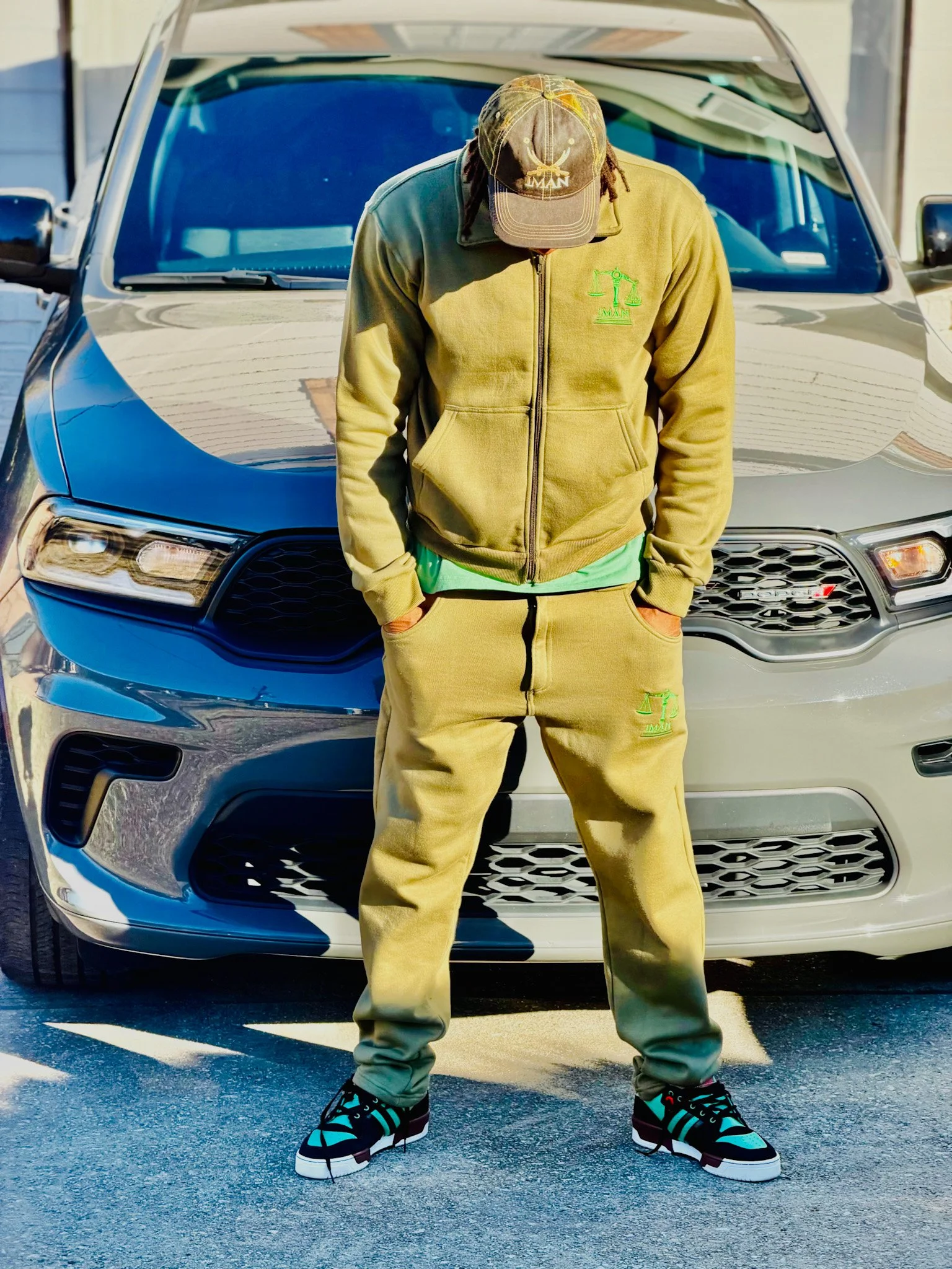 A person dressed in a beige hoodie, pants, and a cap standing in front of a silver car with their head tilted down and hands in pockets.