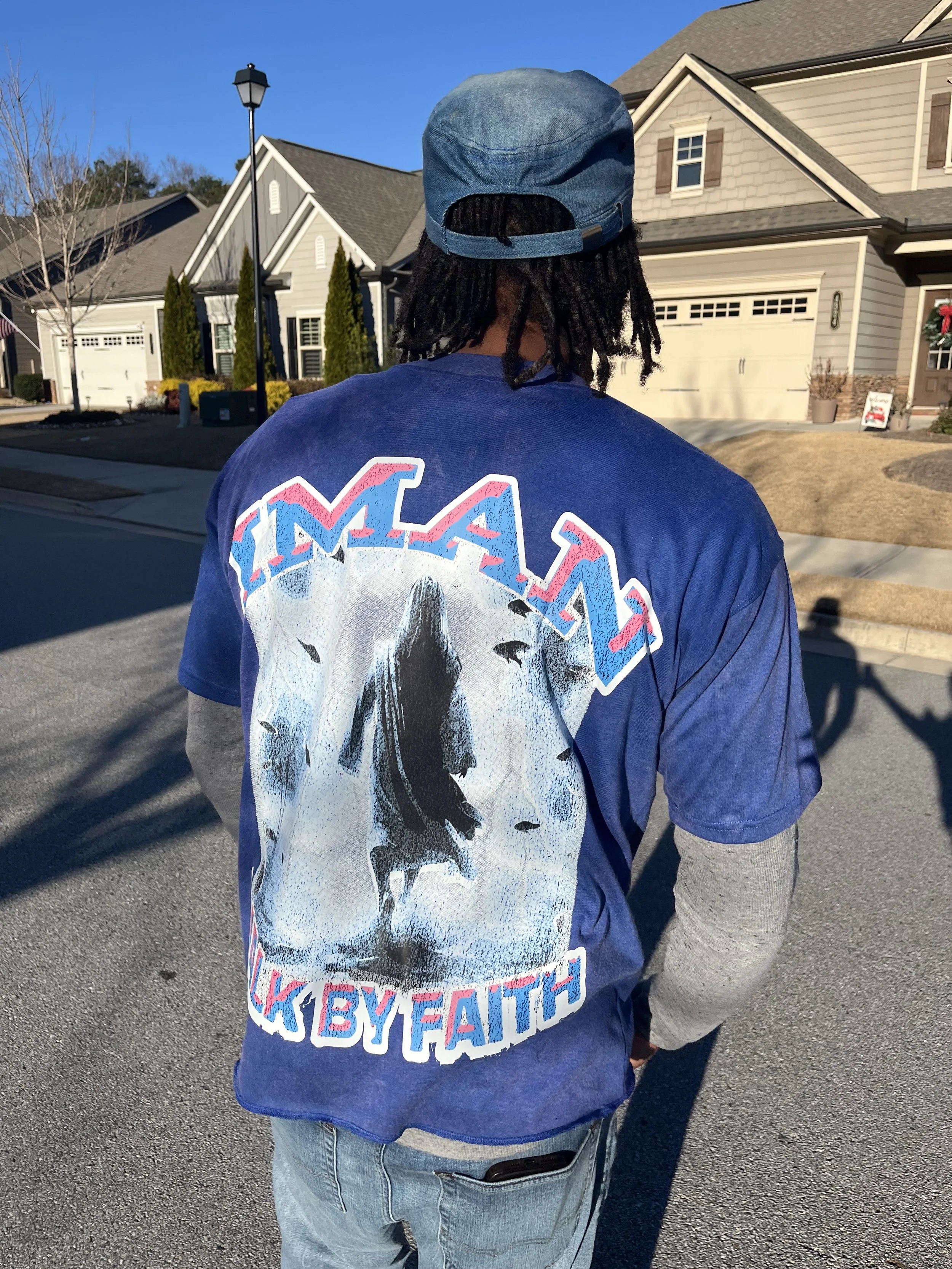 Back view of a person standing on a suburban street, wearing a blue WASPY Faith T-shirt and a denim cap, with houses and a clear blue sky in the background.