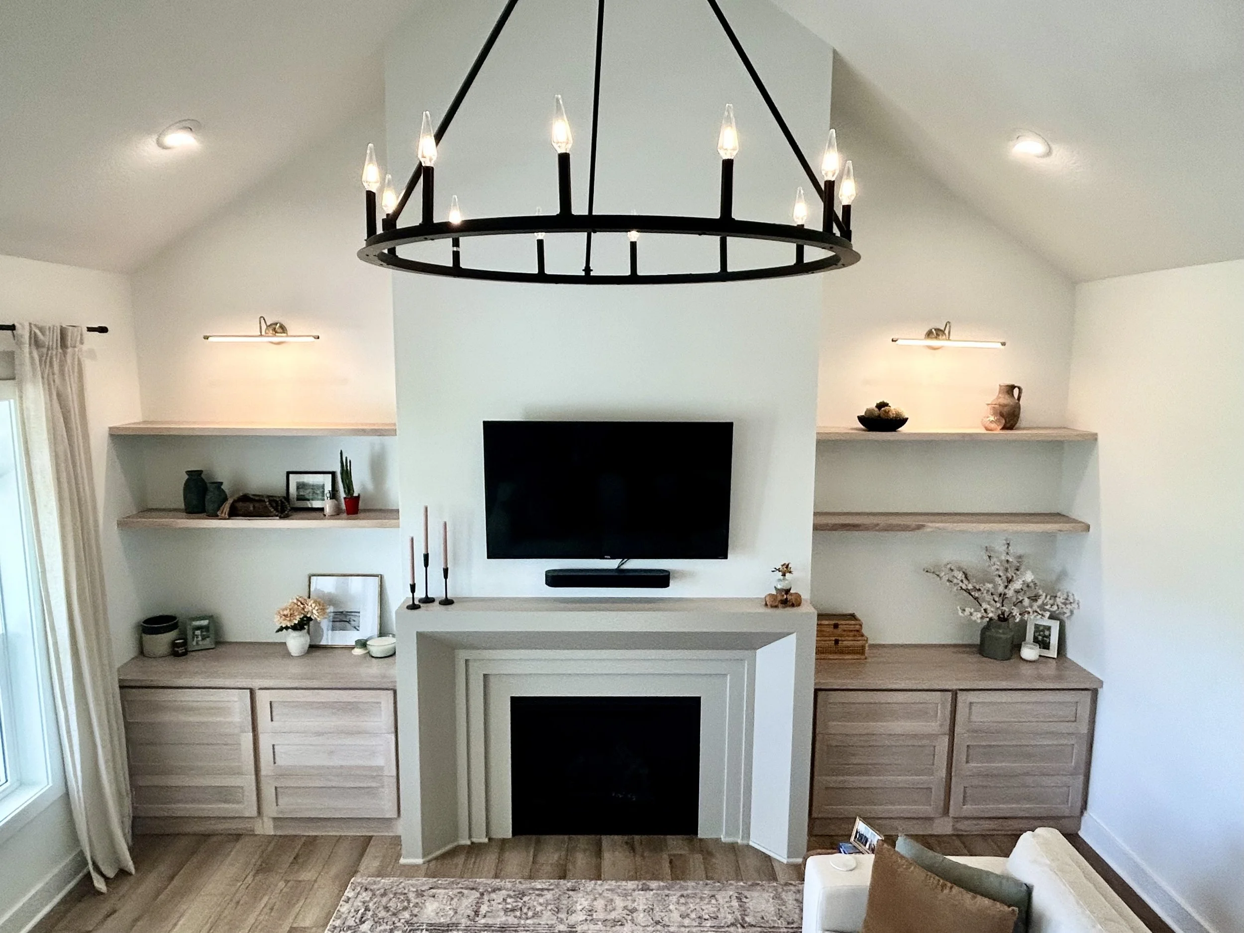 Living room with a high ceiling, central fireplace, mounted television, built-in shelves decorated with vases, picture frames, and plants, and a chandelier hanging from the ceiling.