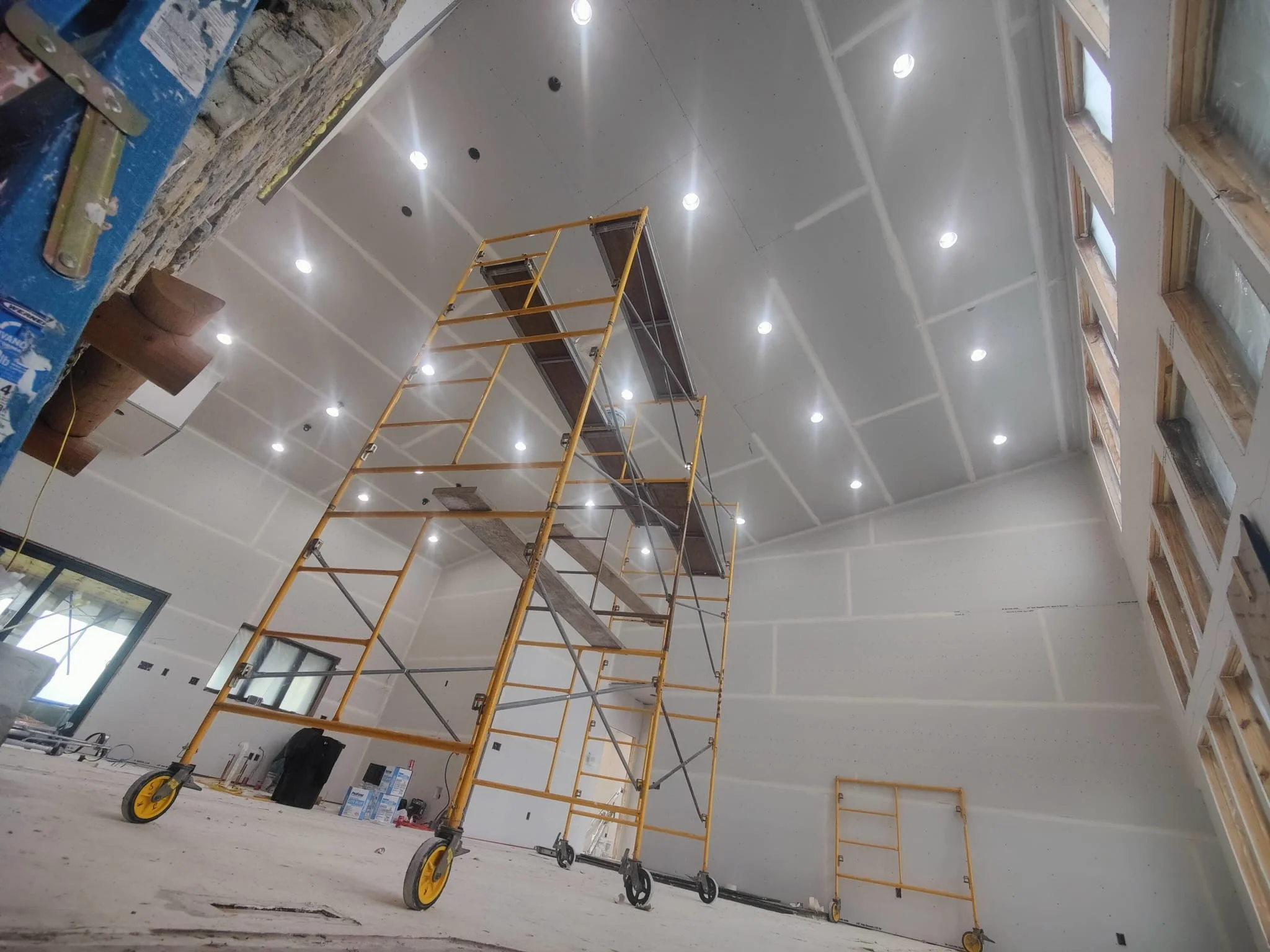 Construction scaffolding inside a large room with unfinished walls and ceiling, multiple ceiling lights, and a row of windows with wooden frames.