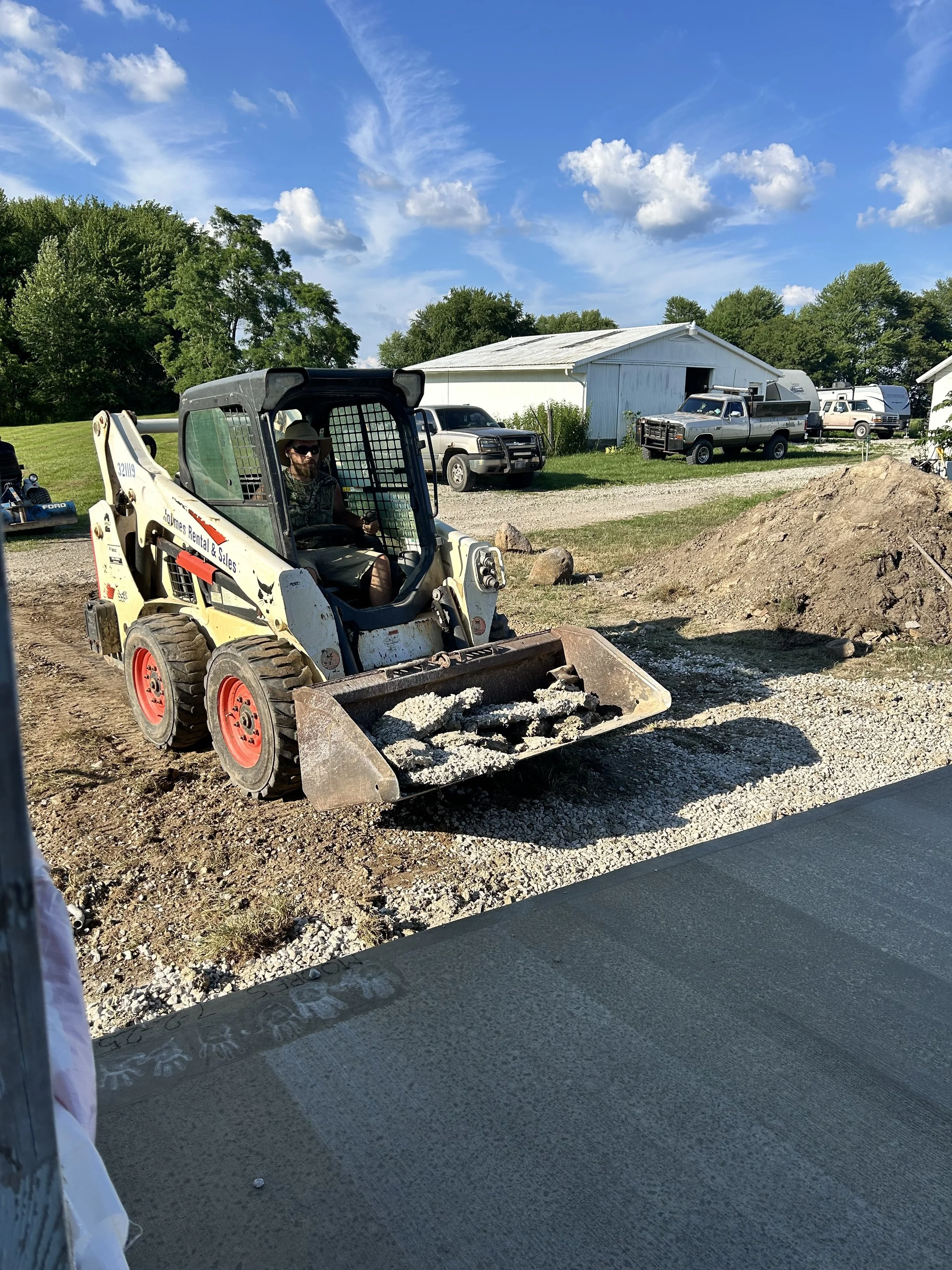 A man operating a small skid-steer loader with a bucket full of gravel on a construction site. The background shows a white shed, several parked trucks, and a blue sky with clouds.