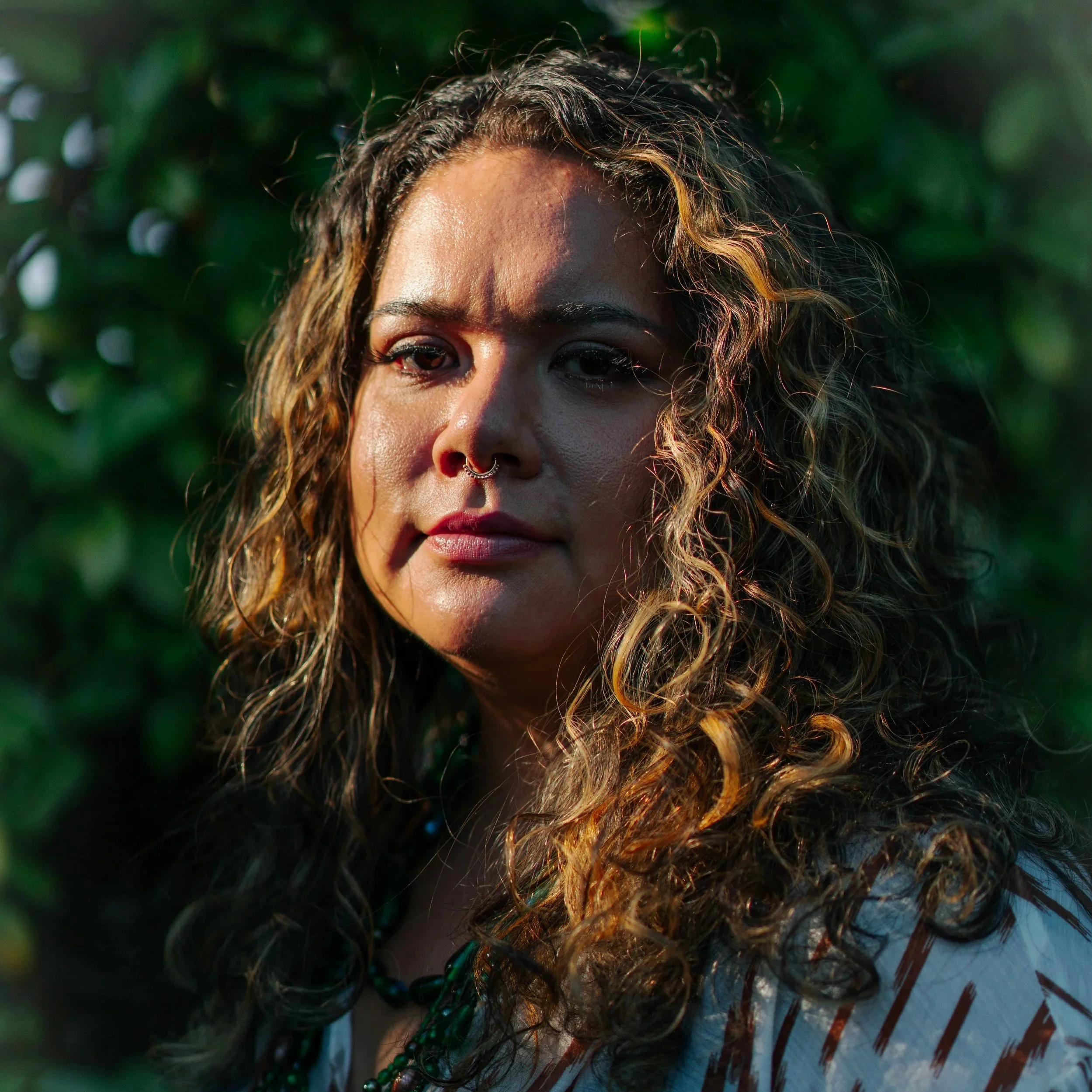A woman with curly hair, a septum piercing, and wearing layered necklaces, standing outdoors with green foliage in the background. Monica Castro headshot and studio photoshoot.
