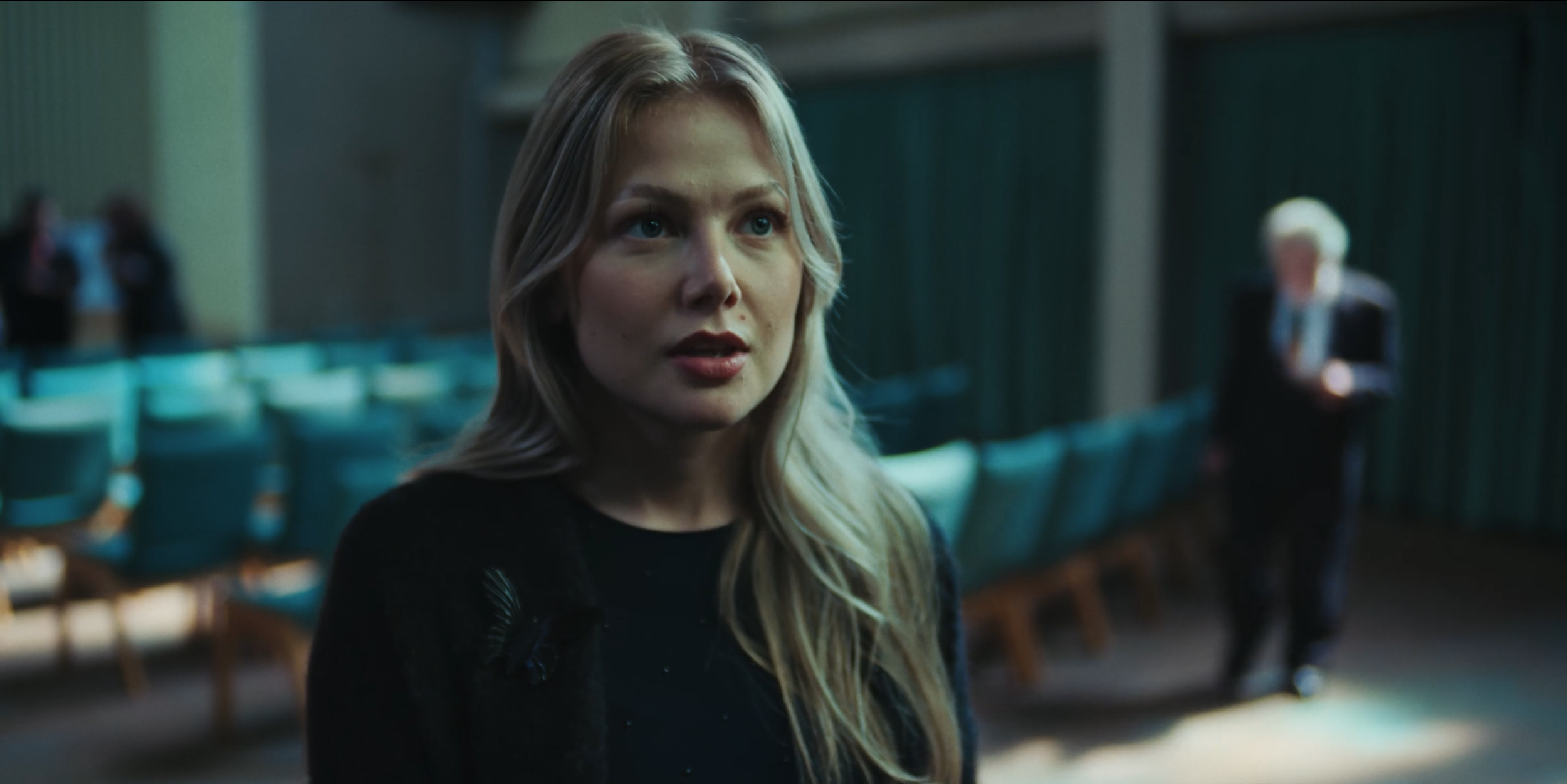 A woman with blonde hair looking surprised or concerned stands in a dimly lit room with teal chairs. In the background, an older woman with white hair is holding a phone, and there are a few other blurred people.