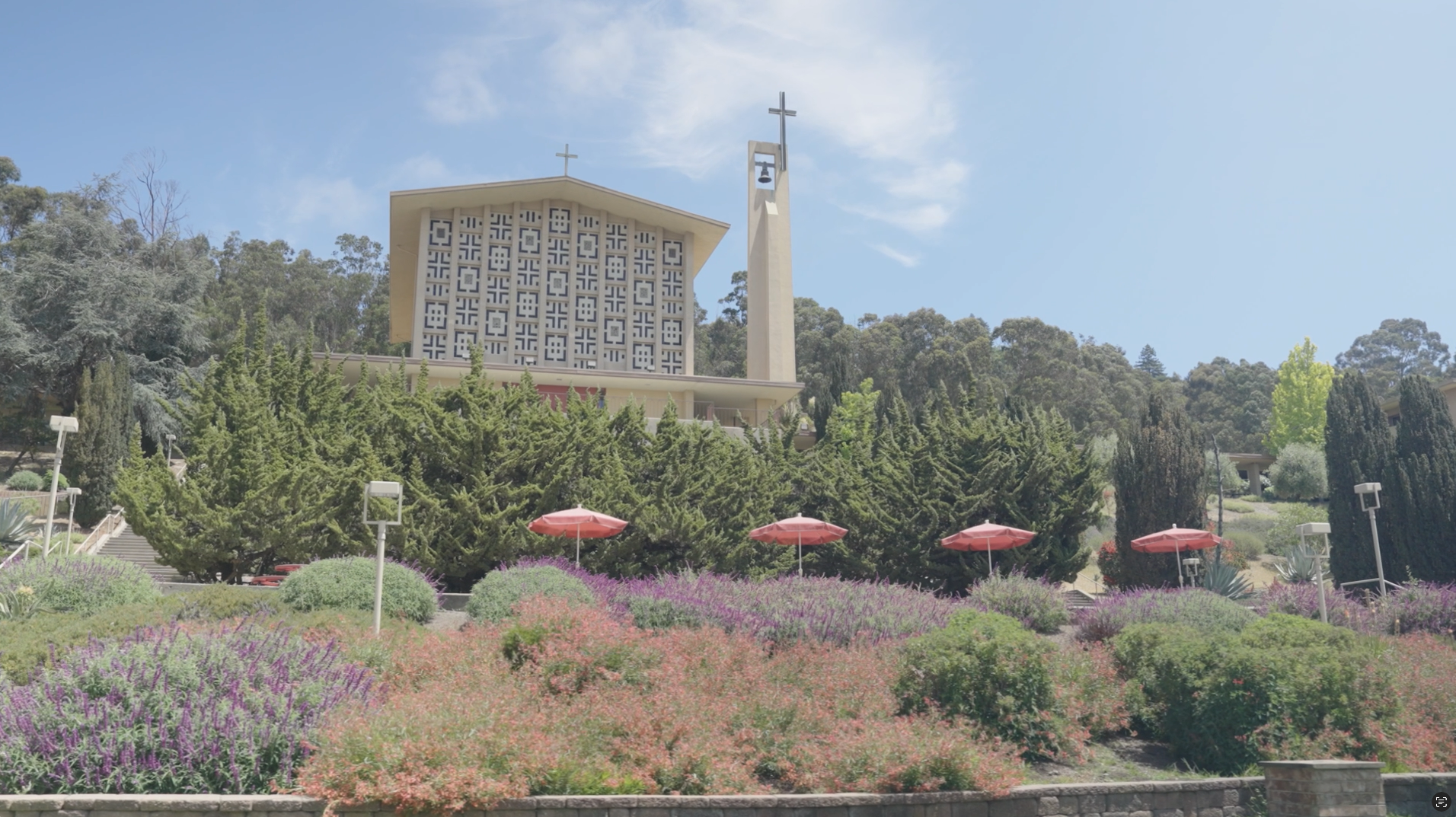 A church with a cross on top, bell tower, modern patterned facade, surrounded by landscaped garden with trees, bushes, colorful flowers, and pink umbrellas under a blue sky.