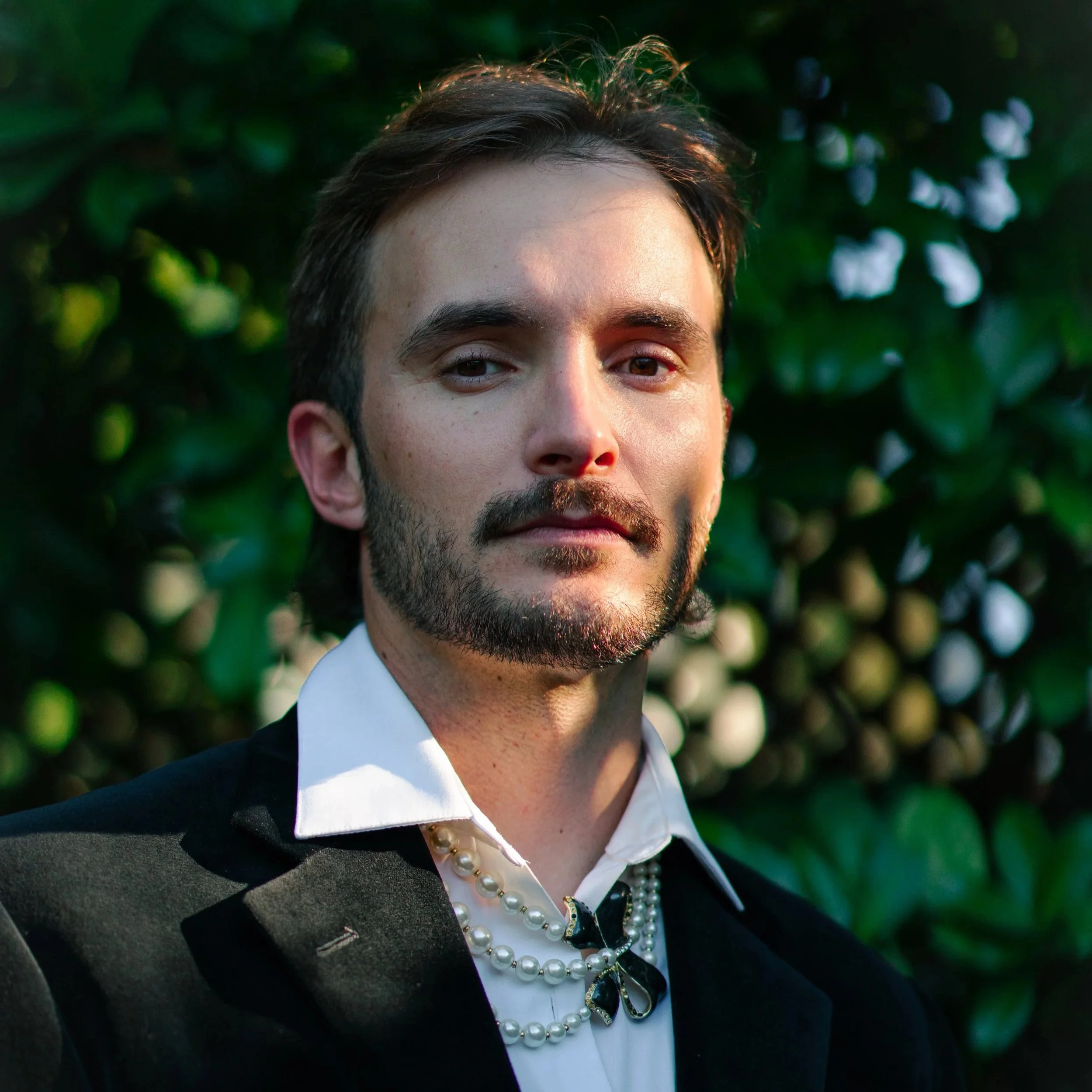 Man in a tuxedo with pearls and a decorative brooch, standing outdoors with green foliage in the background.