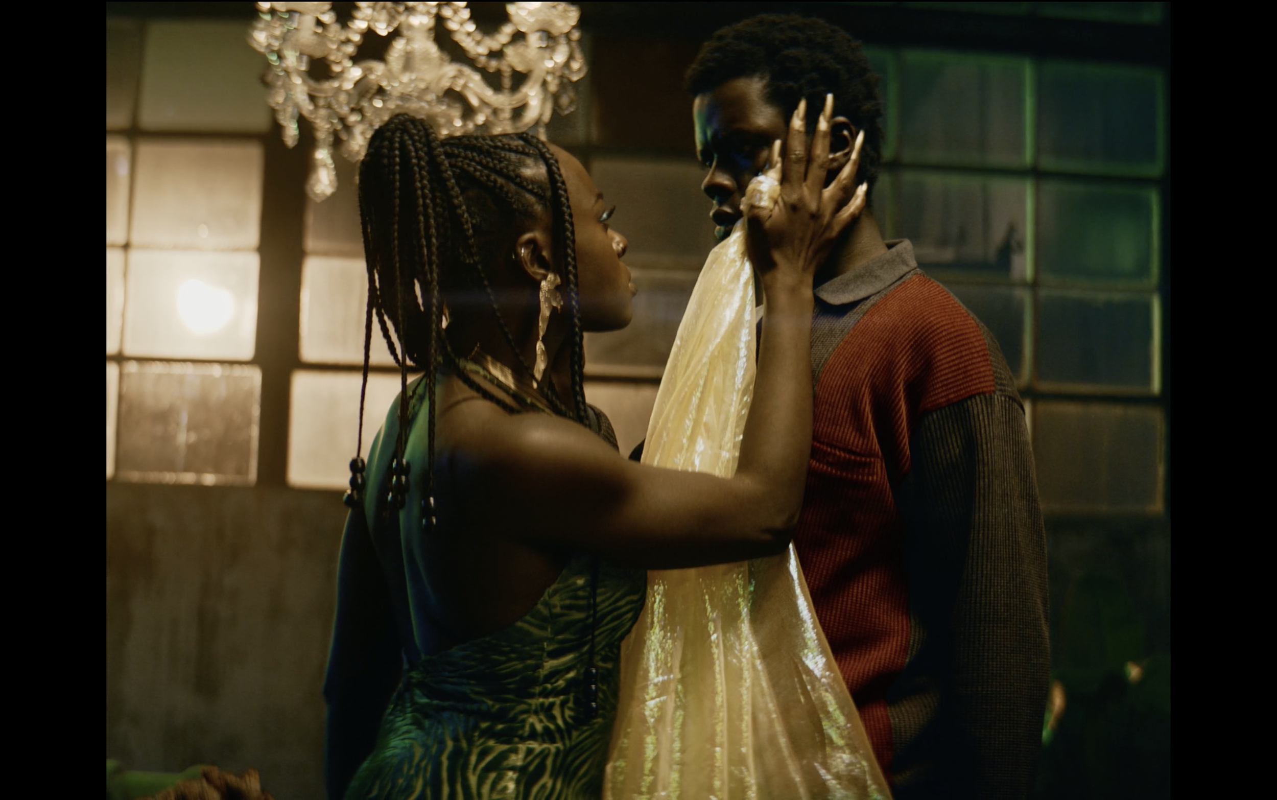 A woman and a man are close together indoors, with the woman touching the man's face delicately. The woman has braided hair adorned with beads and is wearing a patterned dress. The man has short curly hair and is wearing a two-tone sweater. A chandelier hangs above them, and there are glass blocks and warm lighting in the background.