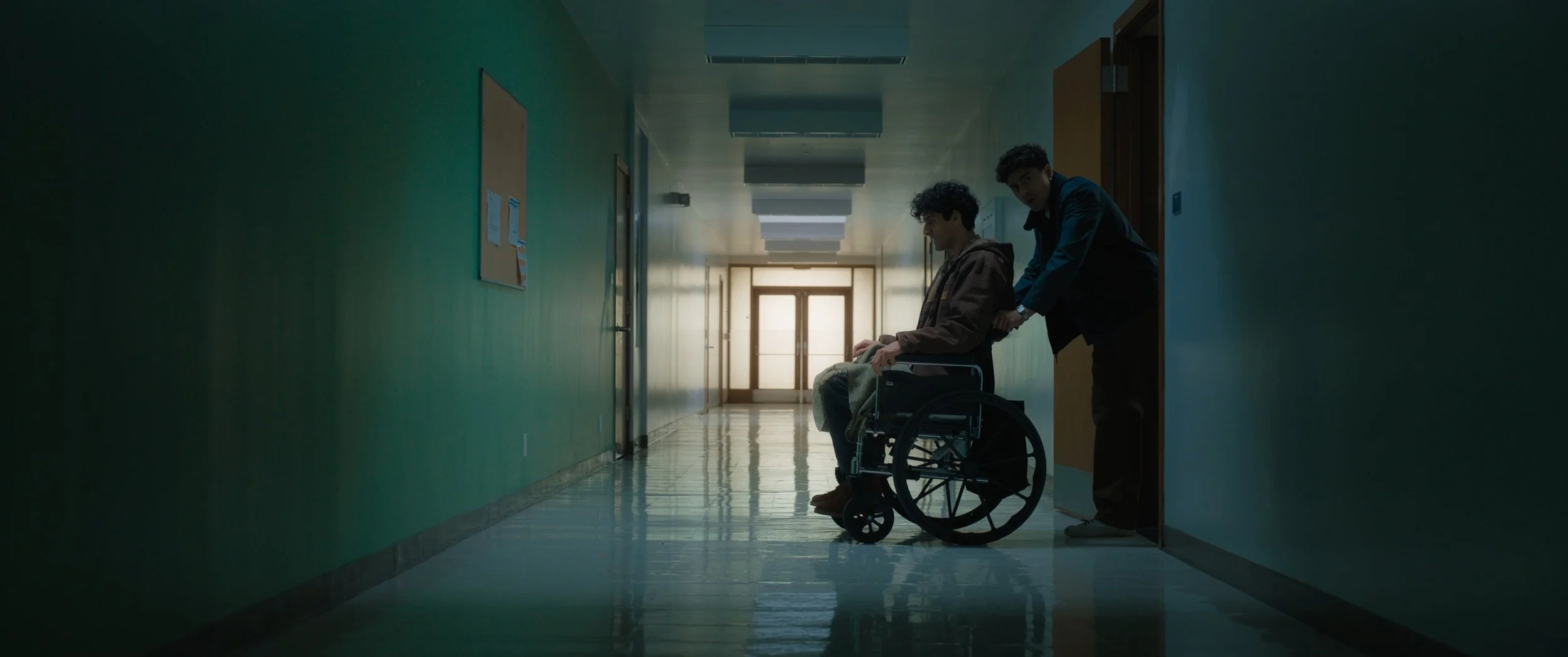 A young person in a brown hoodie sitting in a wheelchair in a hospital corridor, with another person in a black jacket helping them, near an open door at the far end of the hallway.