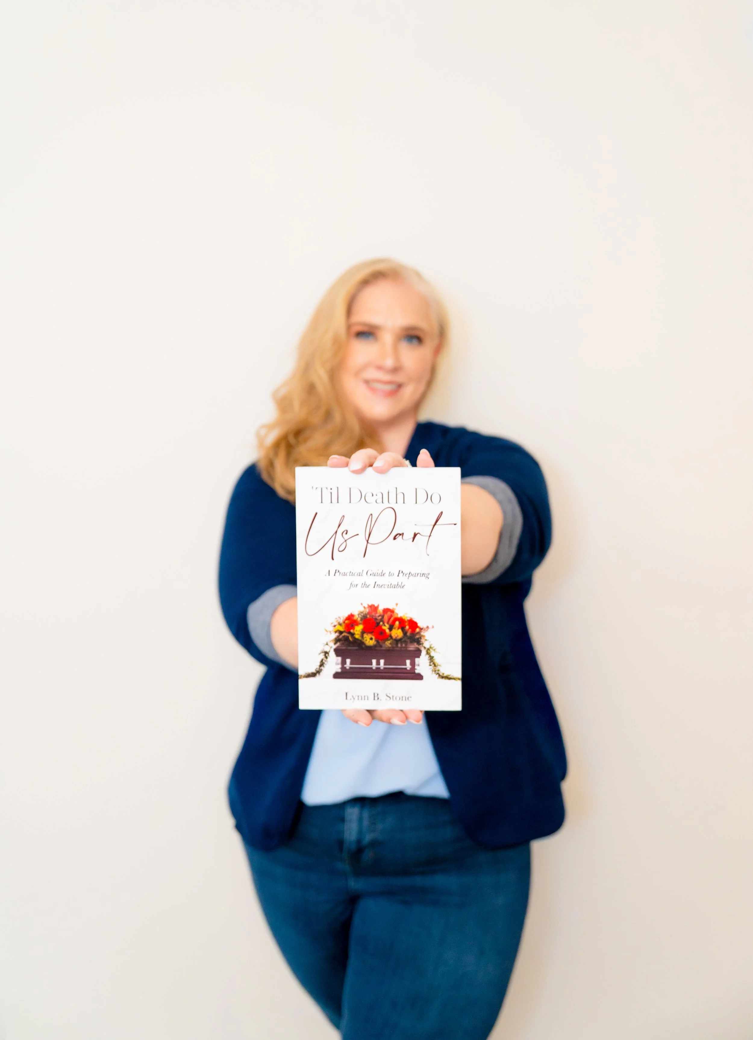 Woman with strawberry-blonde hair wearing a navy blazer and jeans, holding a book titled 'Til Death Do Us Part' in front of her, standing against a plain white wall.