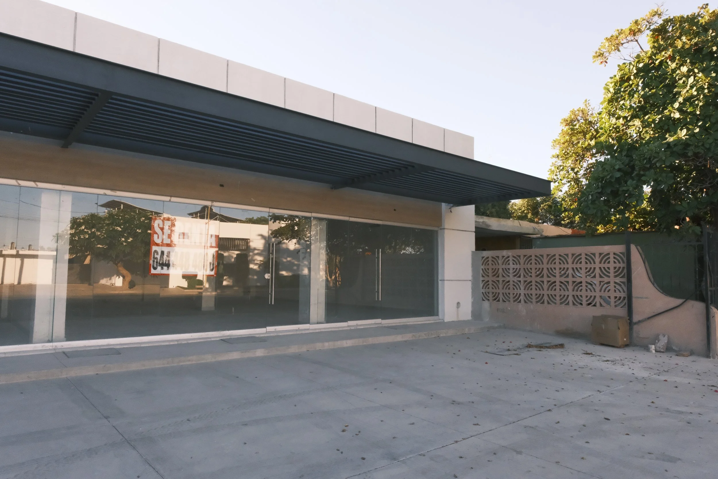 Edificio comercial en venta con fachada de cristal en un barrio residencial, con un letrero que dice "Se vende". Frente de concreto y árbol grande a la derecha.
