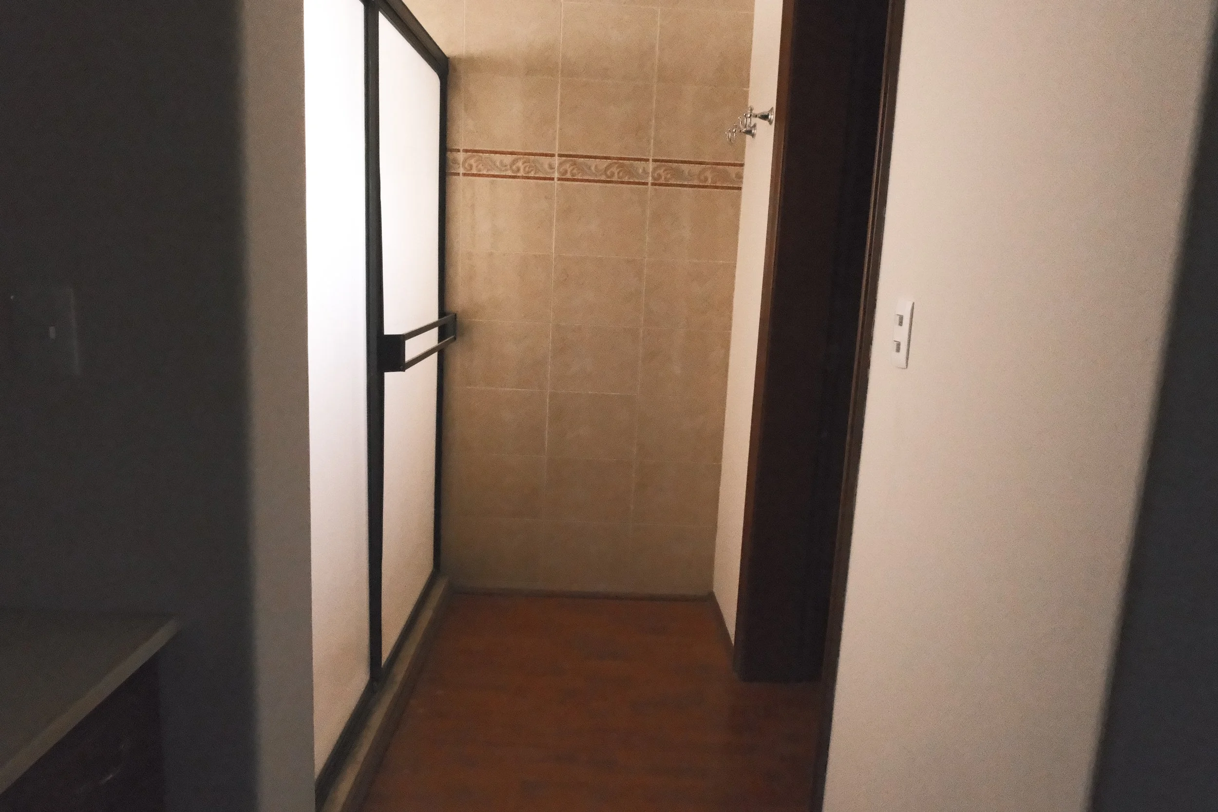 Pasillo con pared de azulejos beige, suelo de madera, y una puerta de aluminio con cristal.