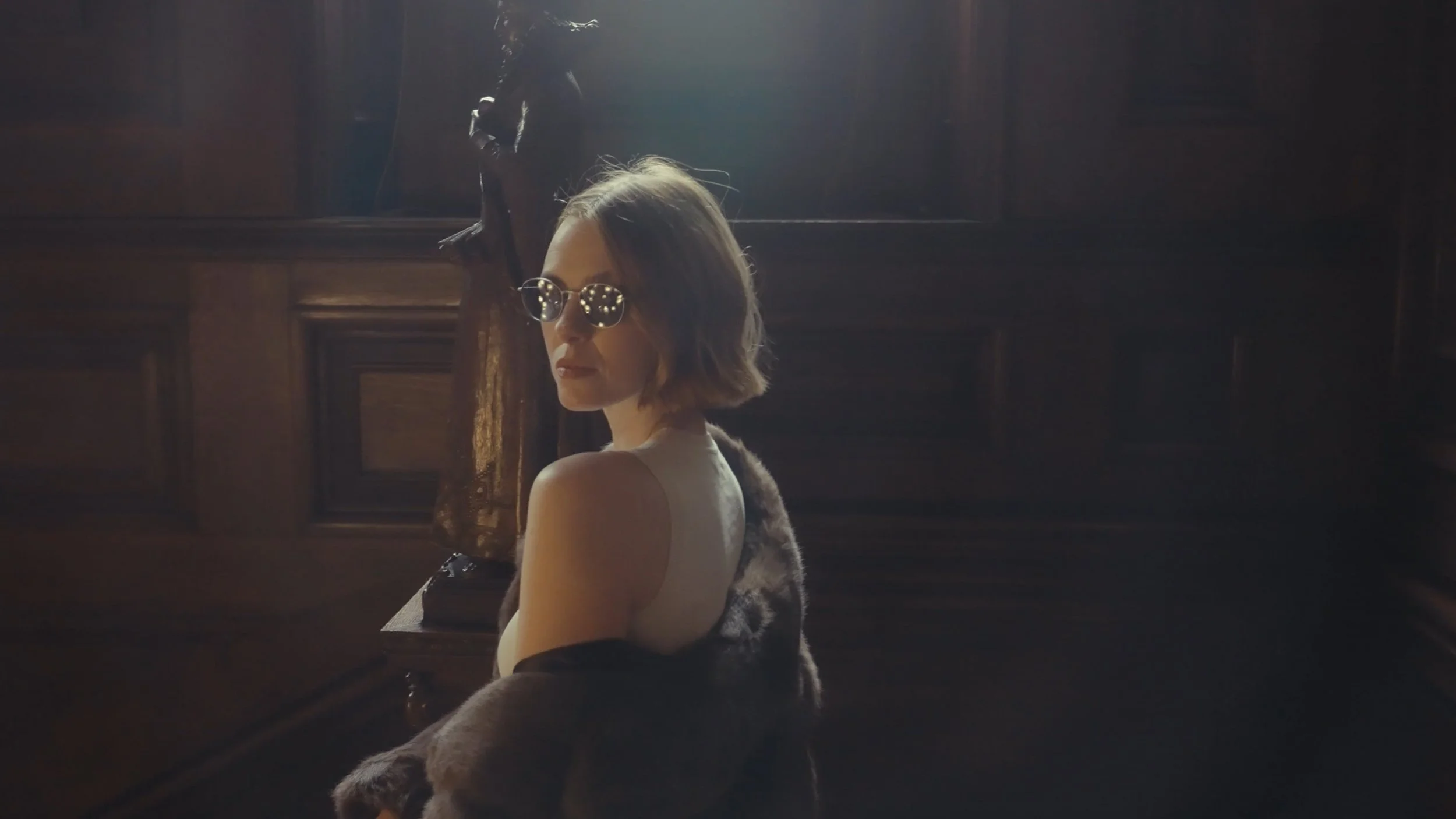 A woman wearing sunglasses and a sleeveless top poses in a dimly lit room with dark wooden paneling, looking over her shoulder.