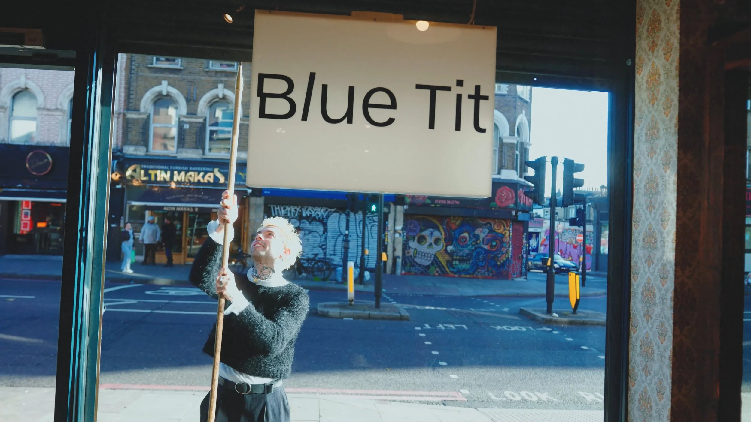 A person with light hair and tattoos, wearing a black sweater and black pants, holding a sign that says 'Blue Tit' outside a building with graffiti art in the background.