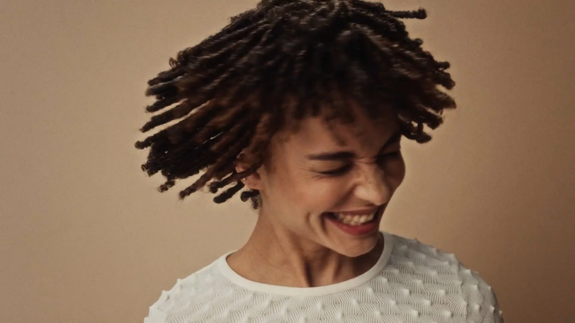 A woman with short, twisted dreadlocks smiling and looking to her left against a beige wall.