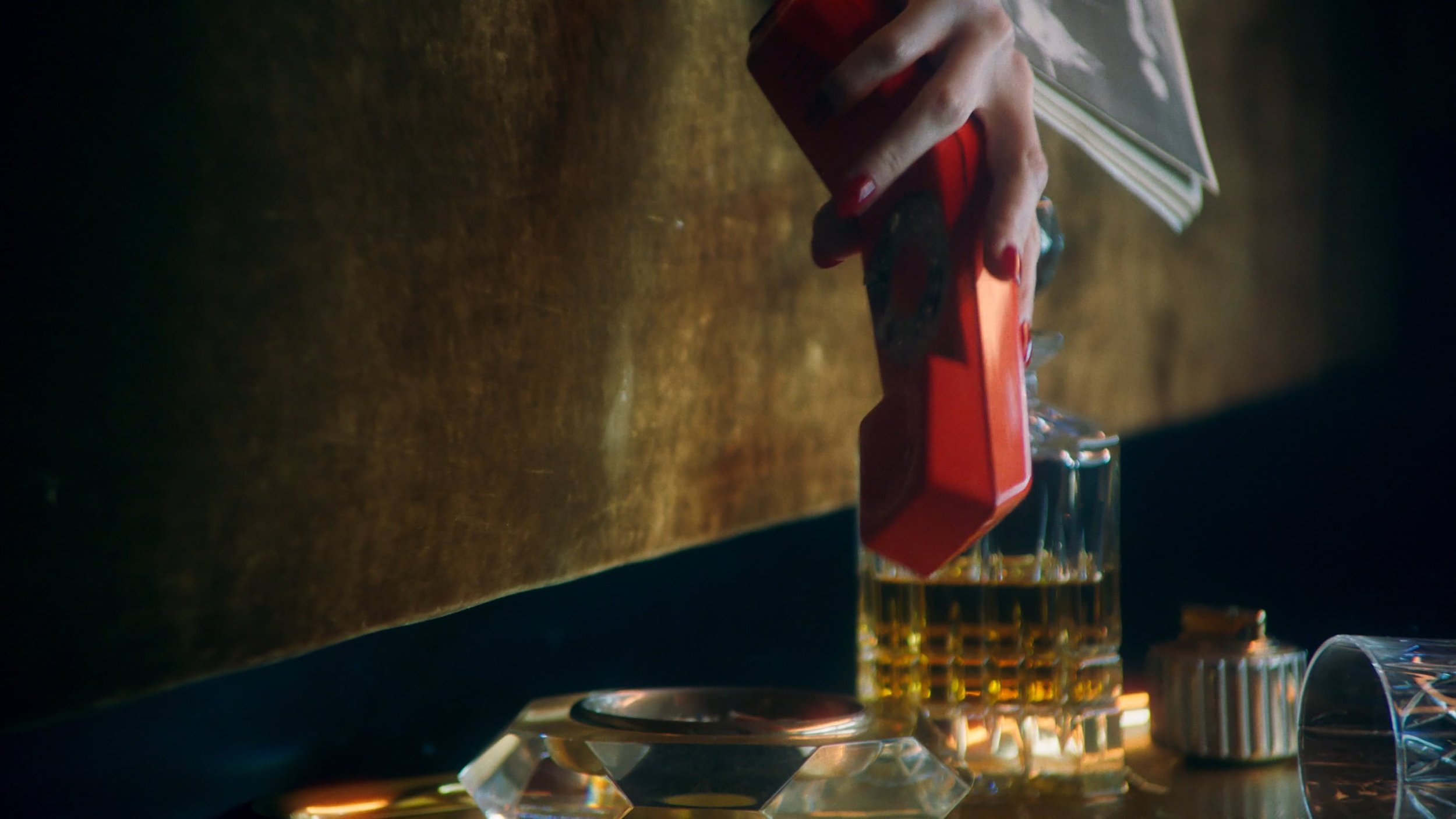 A hand pouring a red liquid from a bottle into a small glass container, with other glasses partially visible on a wooden surface.
