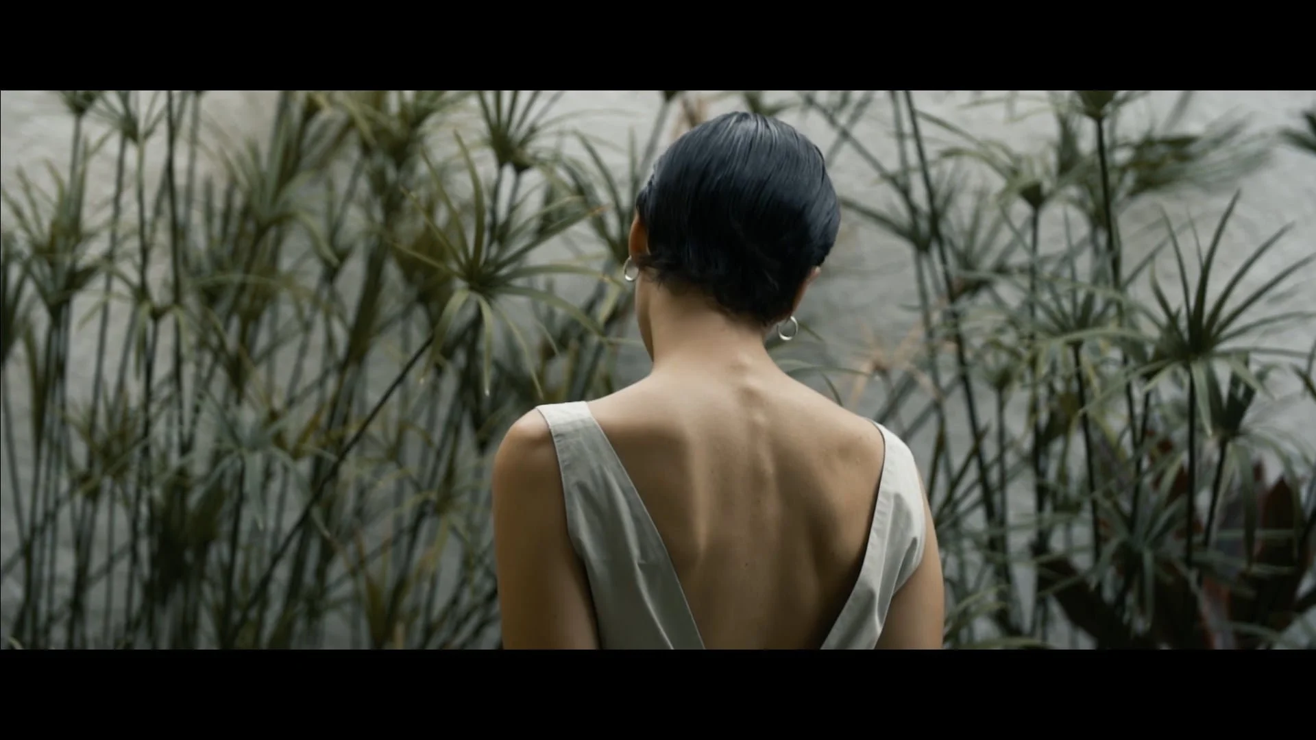 A woman with short black hair wearing a white dress with an open back stands in front of tall green plants.