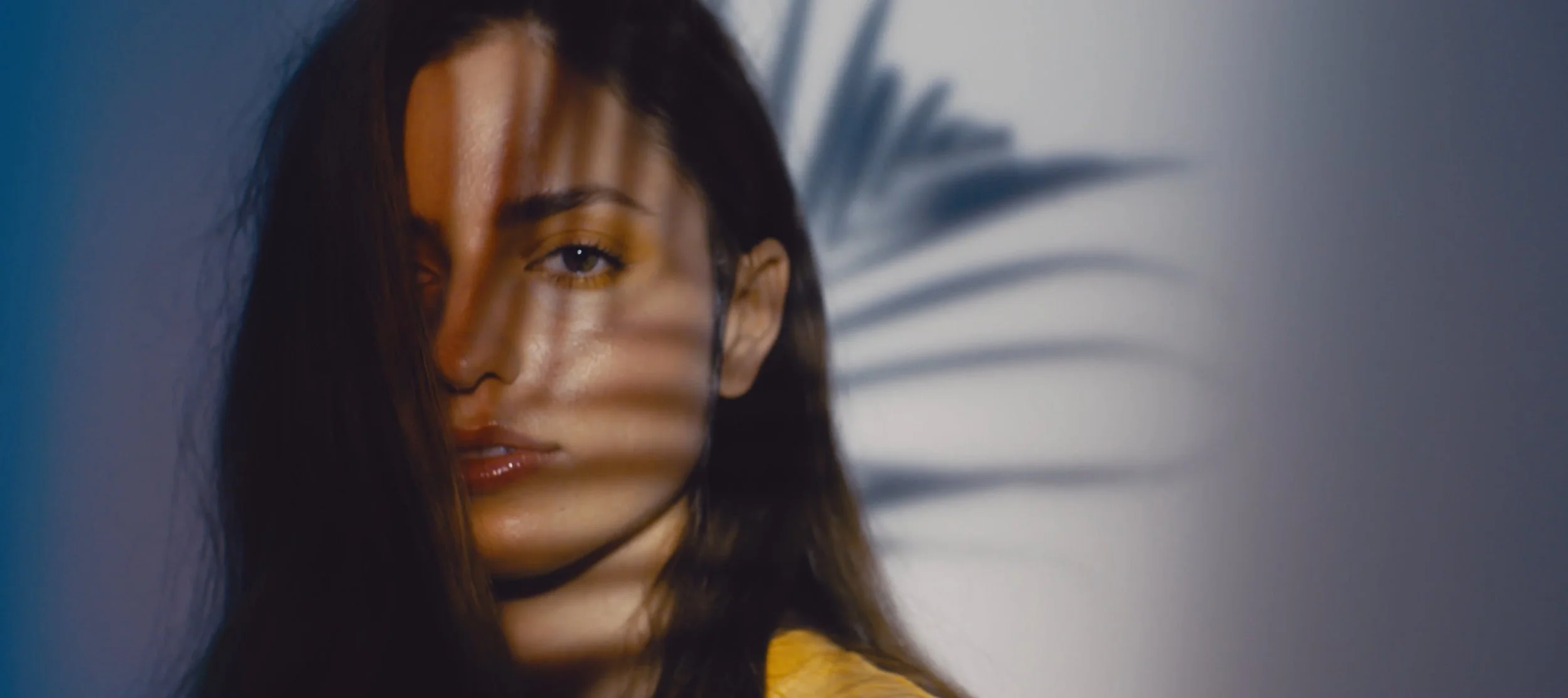 Close-up of a woman with long brown hair, partially obscured by shadows of a plant, looking at the camera.