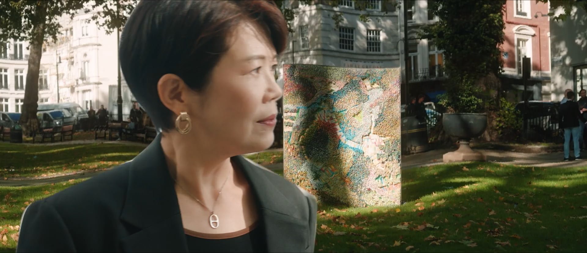 A woman with short dark hair and gold earrings stands outdoors in a park, with paintings displayed behind her. There are trees, benches, and buildings in the background, with several people walking or standing.