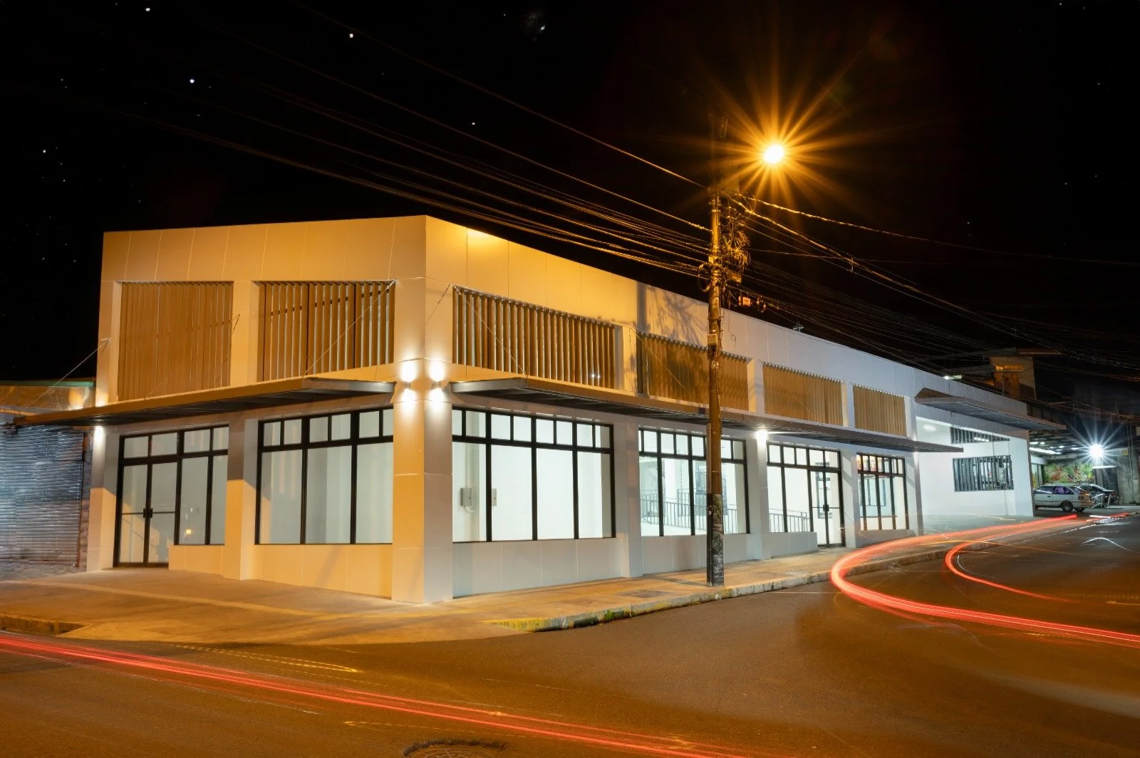 Edificio moderno en esquina iluminado de noche, con ventanales grandes y líneas de movimiento de luz en la calle.