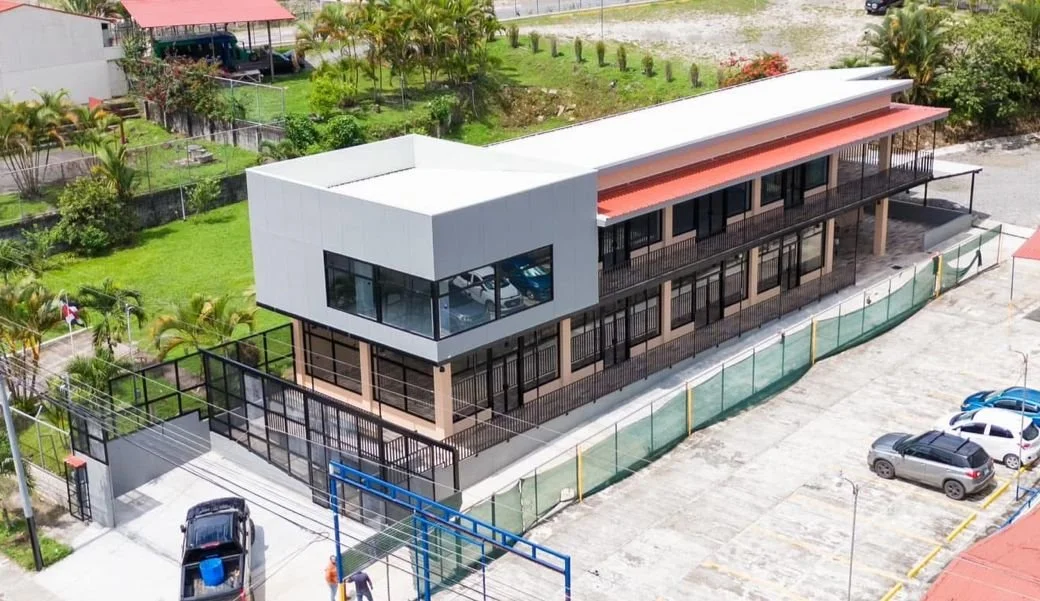 Edificio moderno de dos pisos con ventanas grandes, rodeado de una cerca y estacionamiento con varios automóviles. La estructura tiene un techo rojo y está ubicada en un área verde con jardines.
