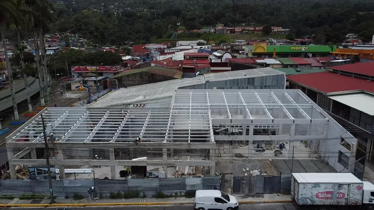 Vista aérea de un sitio de construcción en un área urbana, con estructuras de techos metálicos en proceso y edificios residenciales alrededor.
