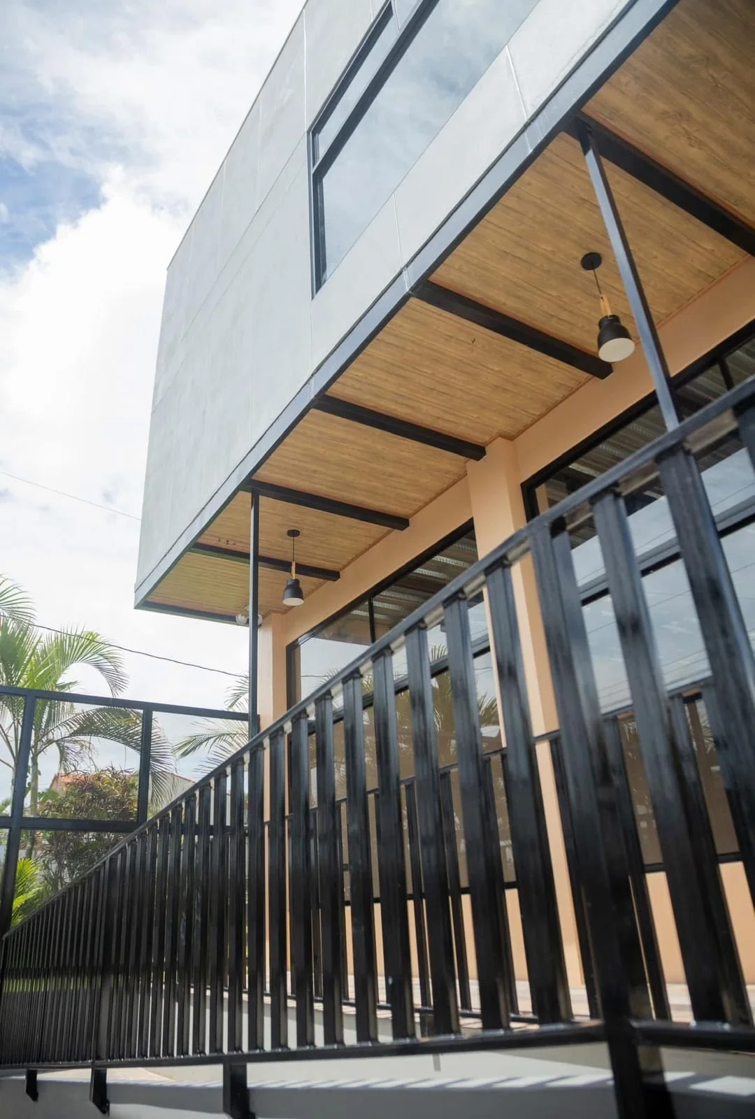 Fachada de un edificio moderno con estructura de metal negro, techo de madera, grandes ventanas y una baranda. Al fondo, un cielo parcialmente nublado y hojas de palmeras.