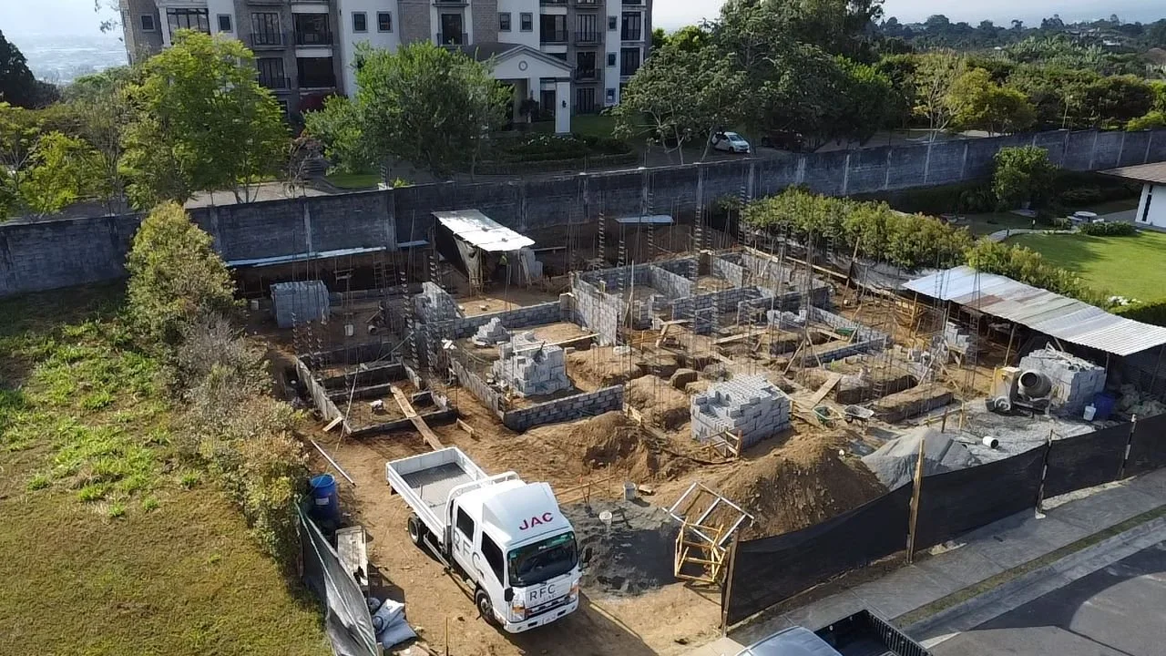 Sitio de construcción con estructuras de concreto, materiales de obra y un camión blanco. Edificio residencial y áreas verdes al fondo.