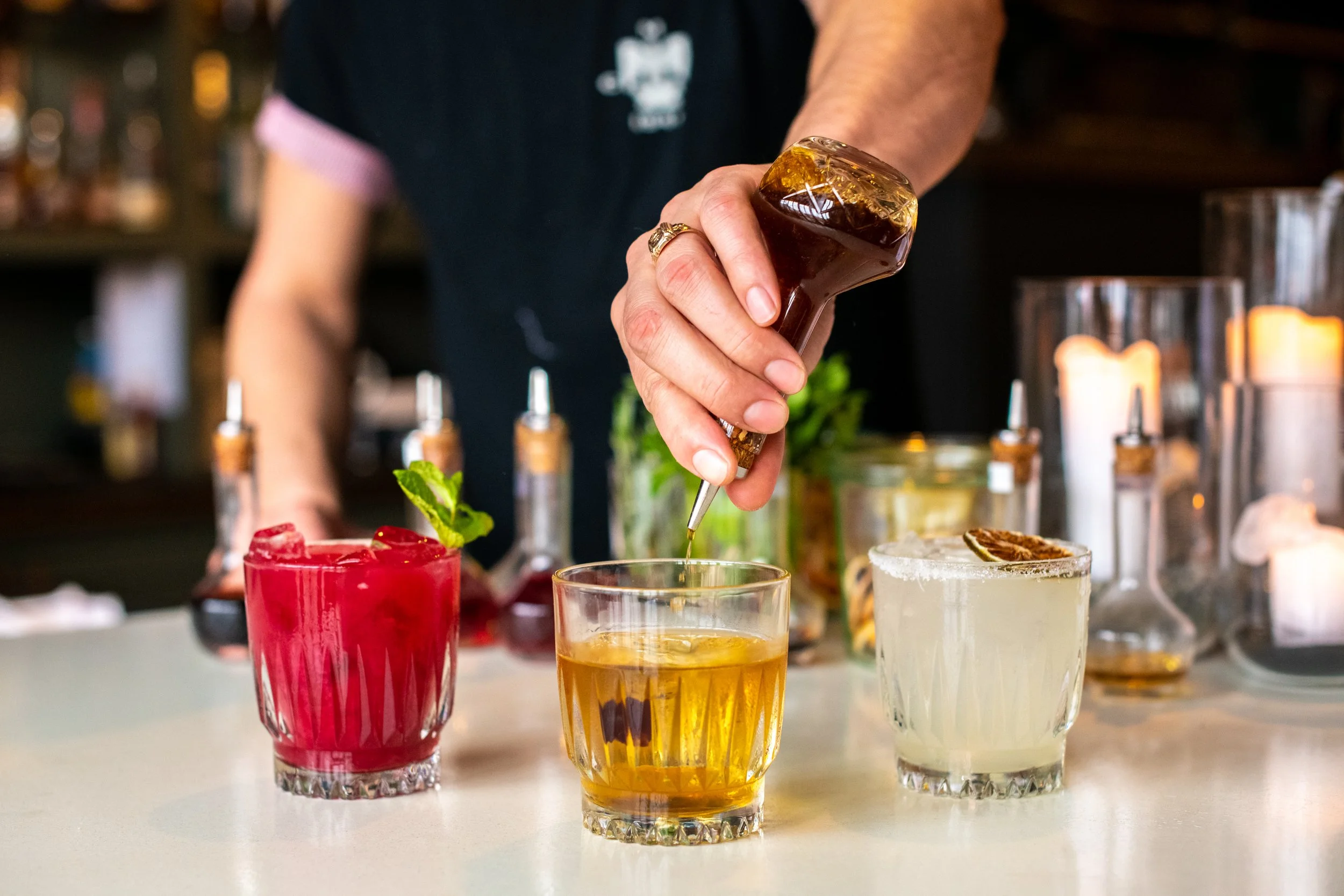 A person adding syrup to a glass of drink among three colorful cocktails on a bar counter.