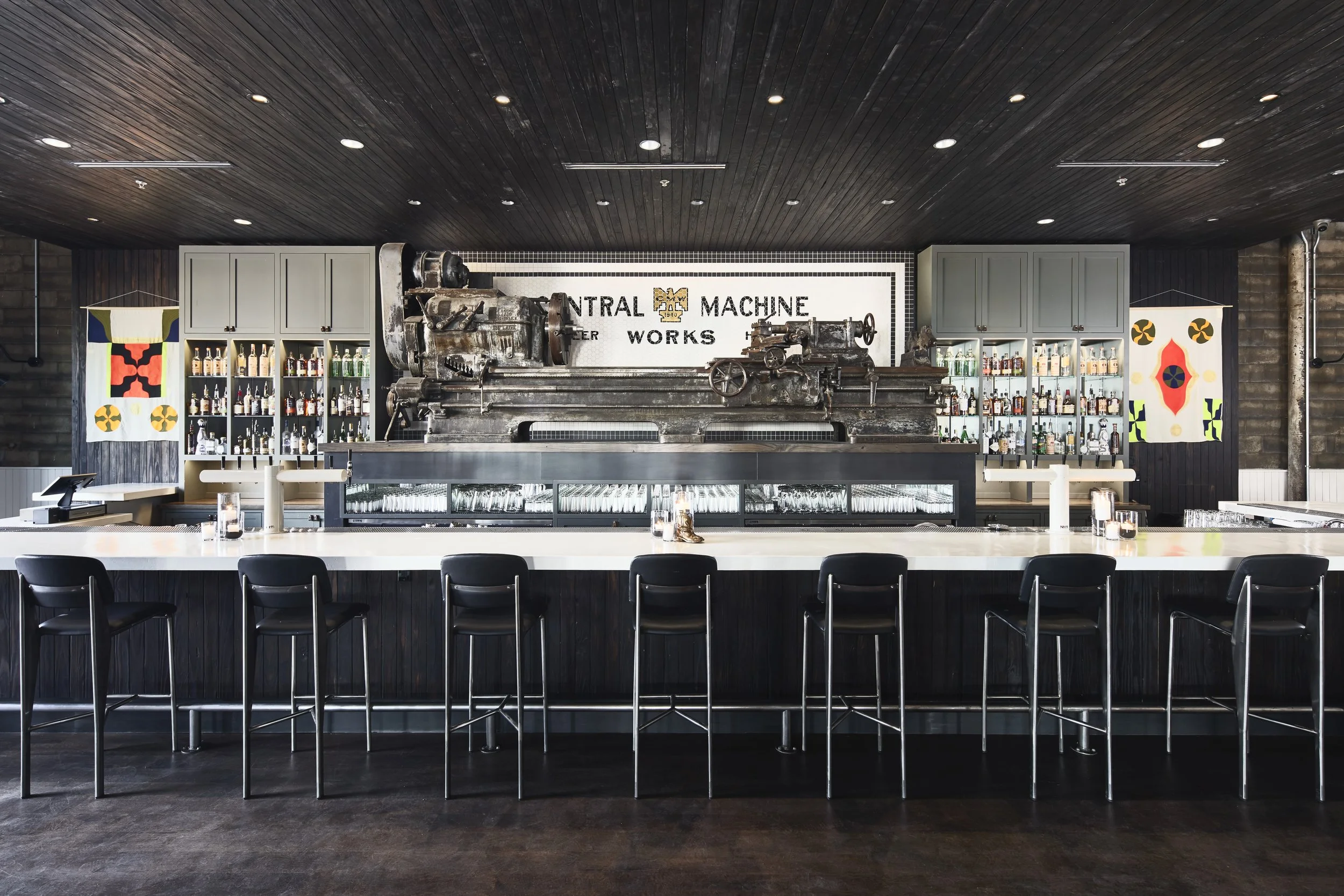 Bar with dark wood ceiling, two shelves of liquor bottles, a vintage machine centerpiece, and colorful abstract banners on each side.