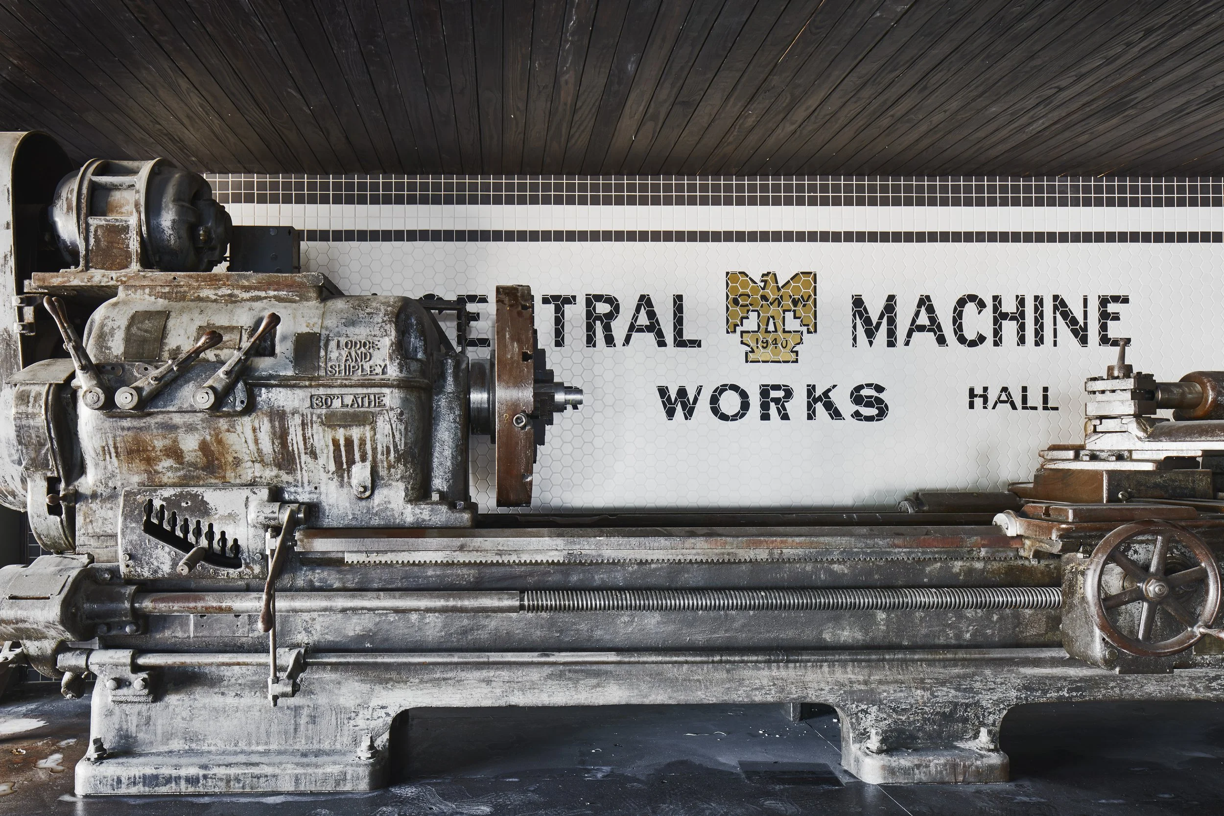An old, rusted industrial machine with various levers and components, placed in front of a white sign that reads 'Central Machine Works Hall' with a logo featuring a honeycomb pattern.