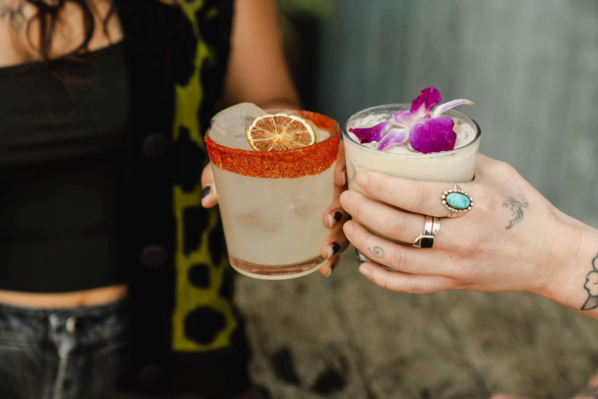 Two women holding cocktails with decorative garnishes, including dried orange slices and purple orchids.