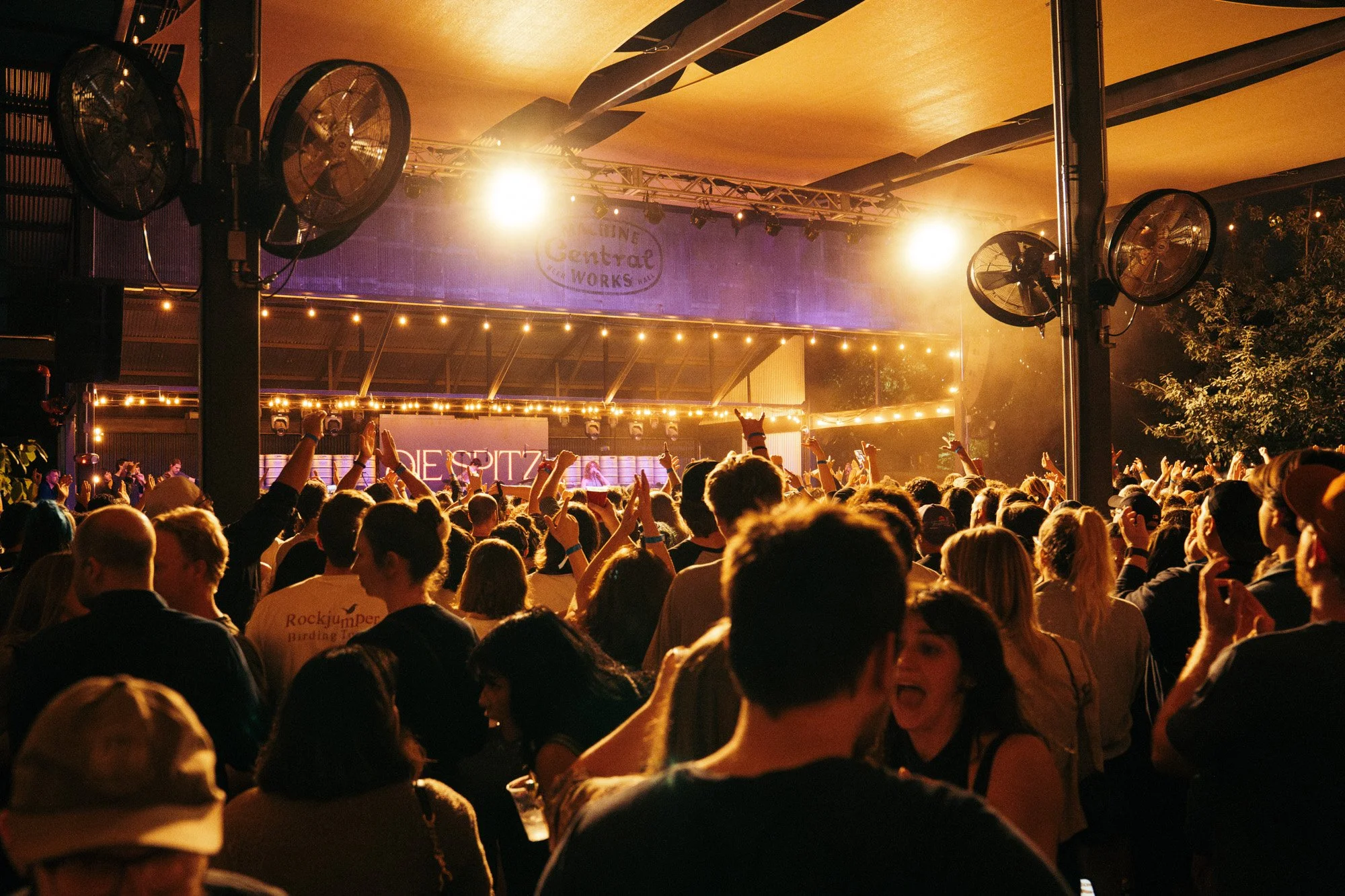 A large crowd of people at night attending an outdoor concert or event. The stage is lit with bright yellow and purple lights, and there are fans mounted on the columns in the foreground. The audience appears to be enjoying the performance, with some
