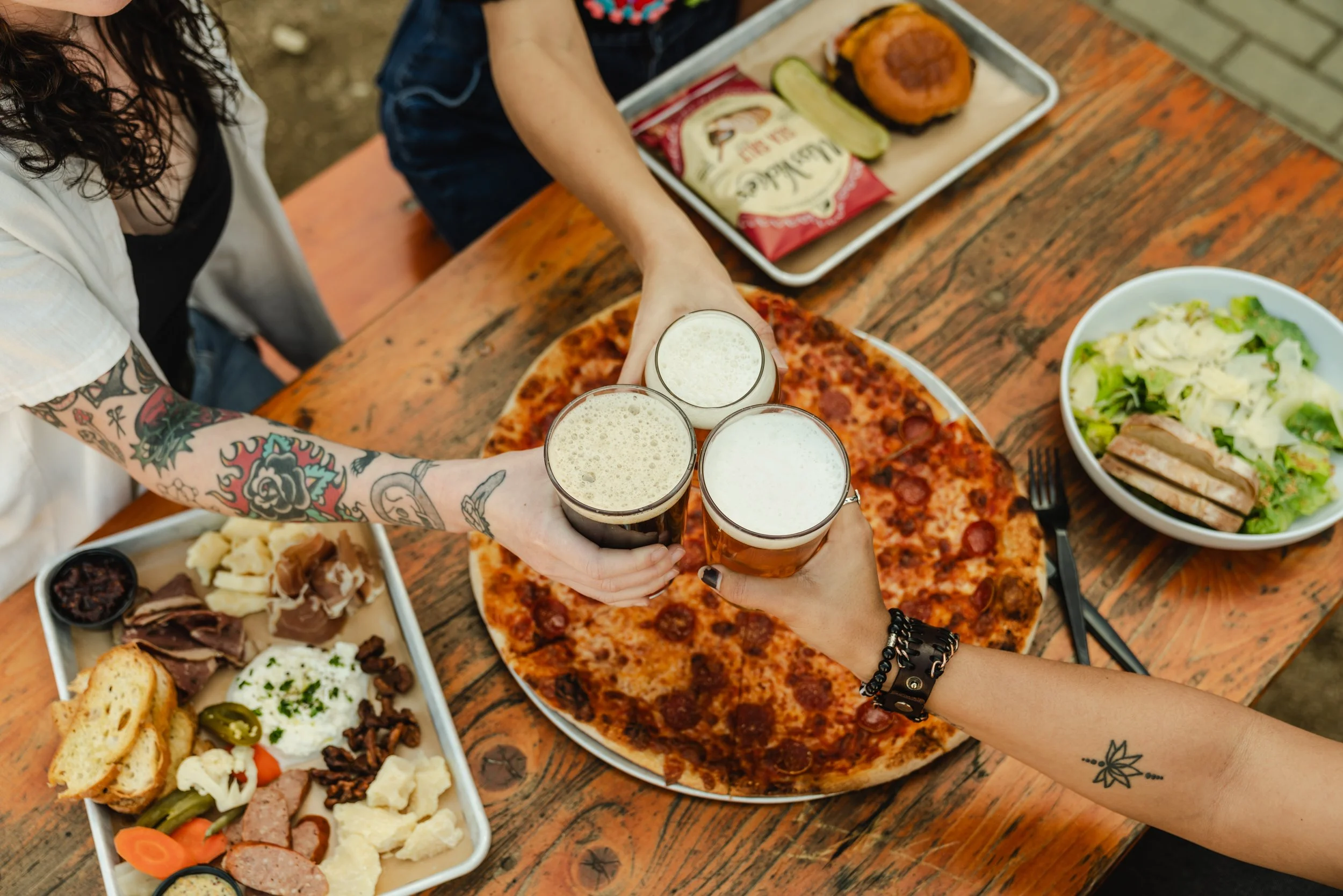 Multiple people clinking glasses of beer over a table with pizza and assorted dishes, including a salad and charcuterie, in a casual dining setting.