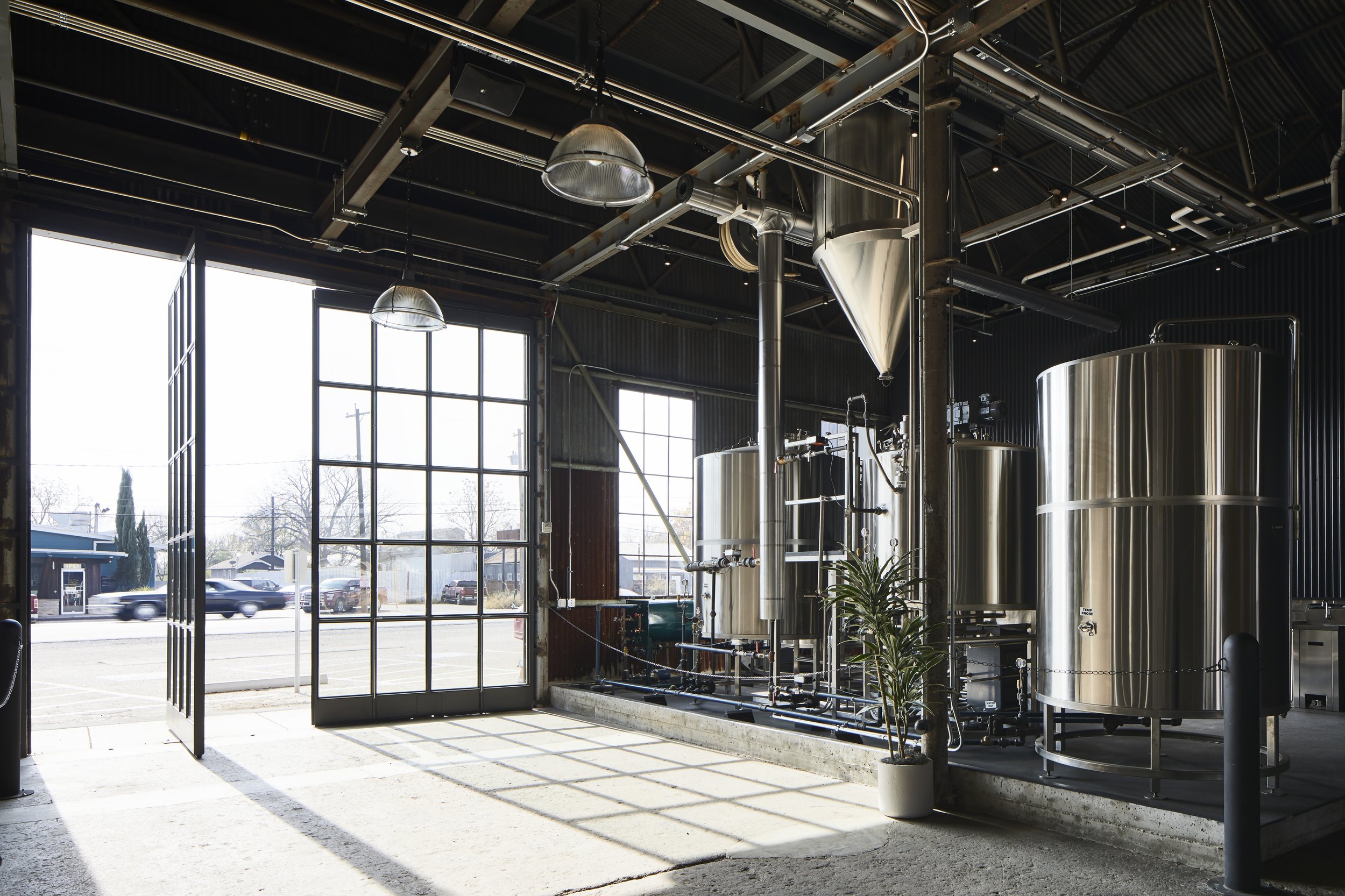 Interior view of a brewery with large stainless steel tanks, pipes, and industrial equipment, with large windows letting in natural light.