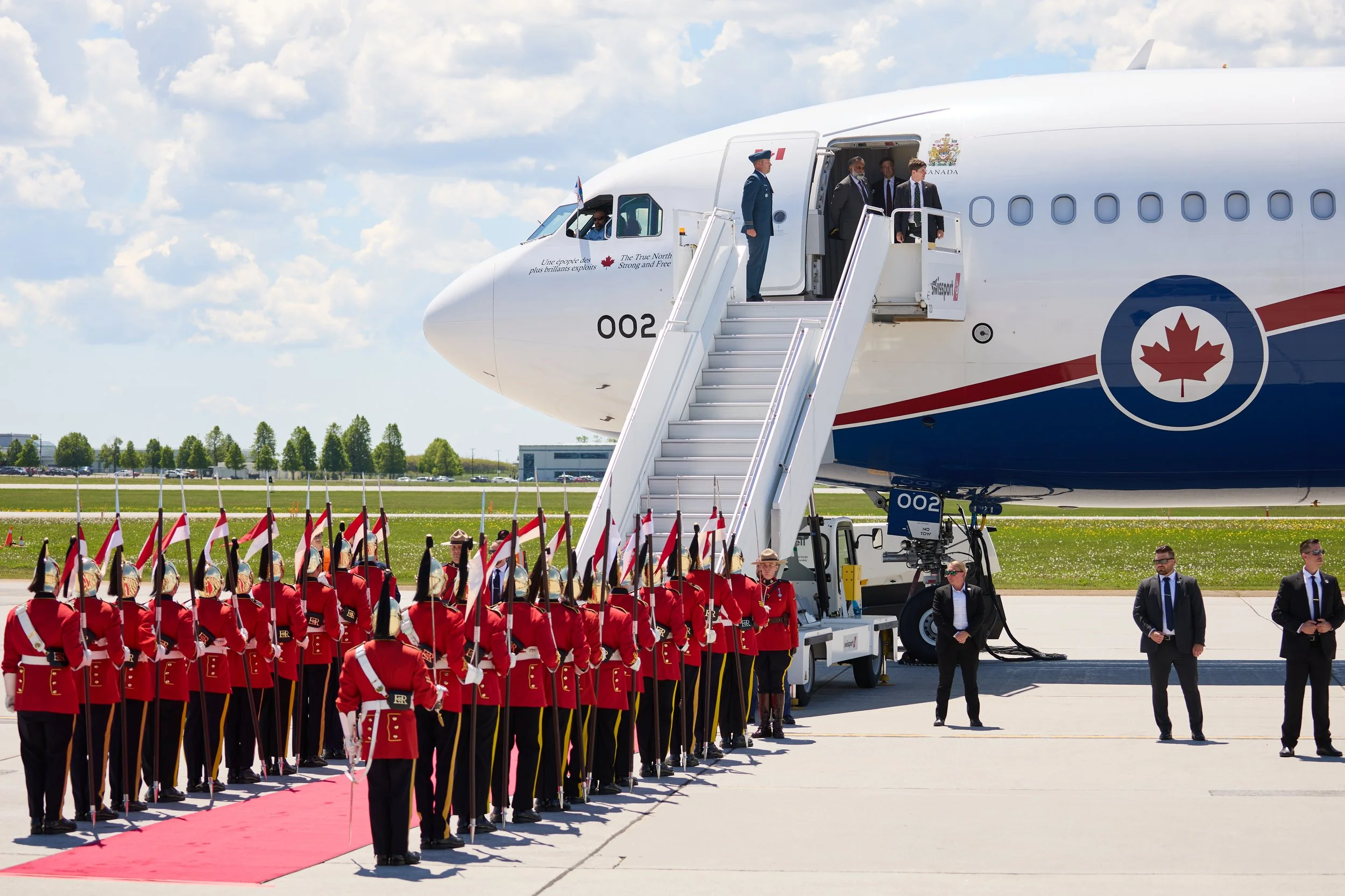Military personnel in red uniforms standing in formation beside a red carpet near a large airplane with a Canadian emblem. Passengers are disembarking from the plane via stairs, with security personnel nearby. The scene is set on an open airport tarm