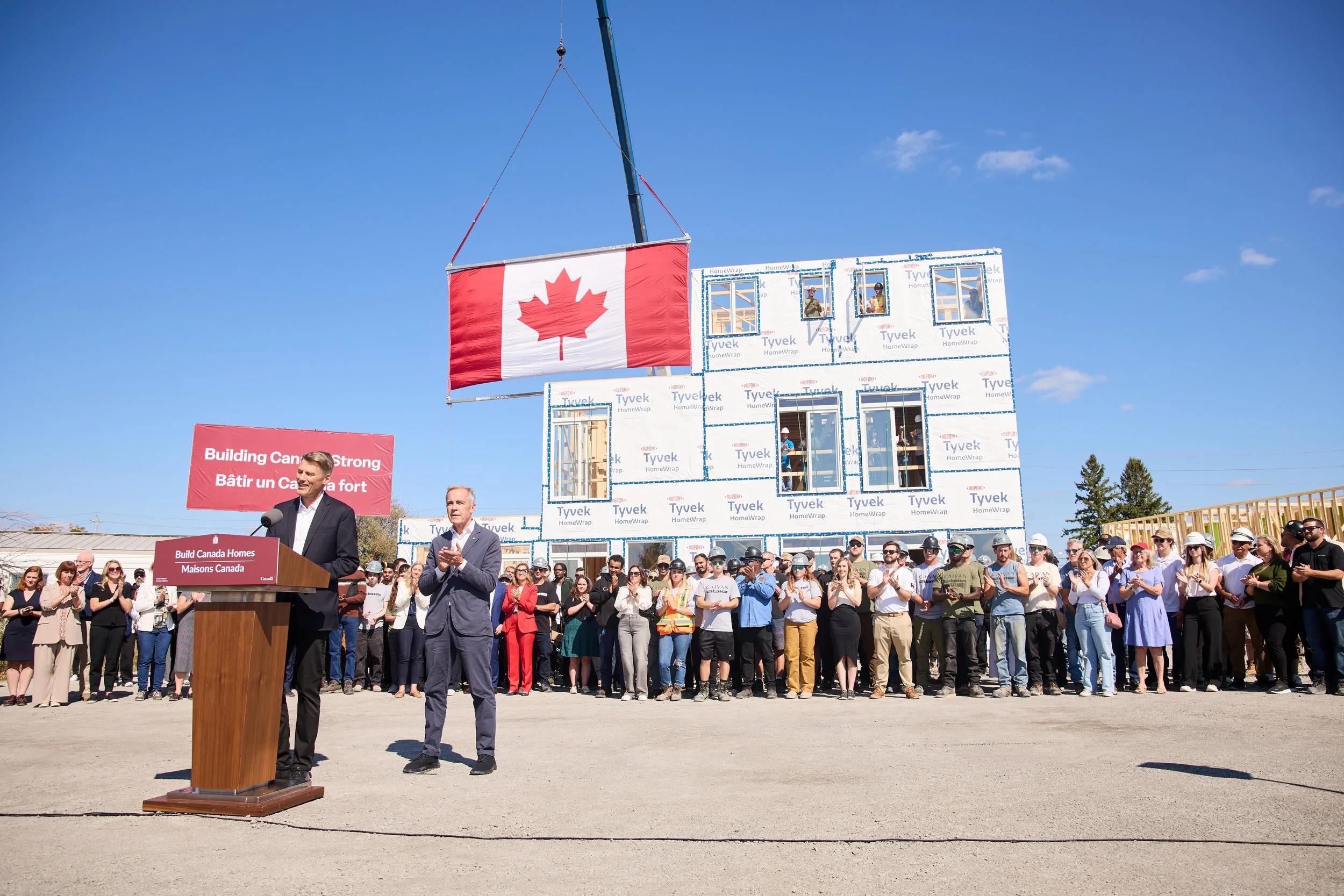 A large group of people attending a groundbreaking ceremony for building homes in Canada. There is a speaker at a podium with a sign that reads 'Build Canada Homes.' Behind the crowd, a house under construction with Tyvek exterior wrap and windows is