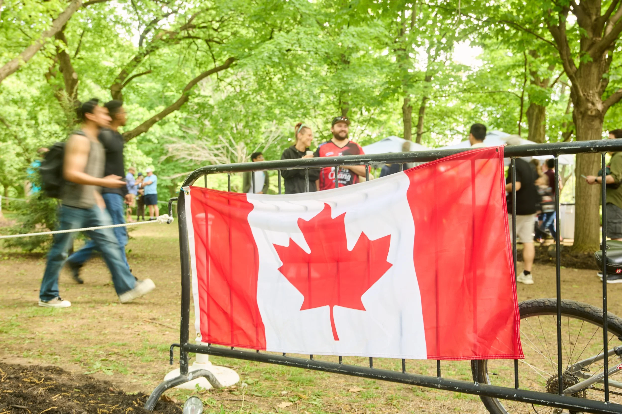 People gathering outdoors in a park with green trees, a Canadian flag draped over a black barricade, and a bicycle partially visible on the right.