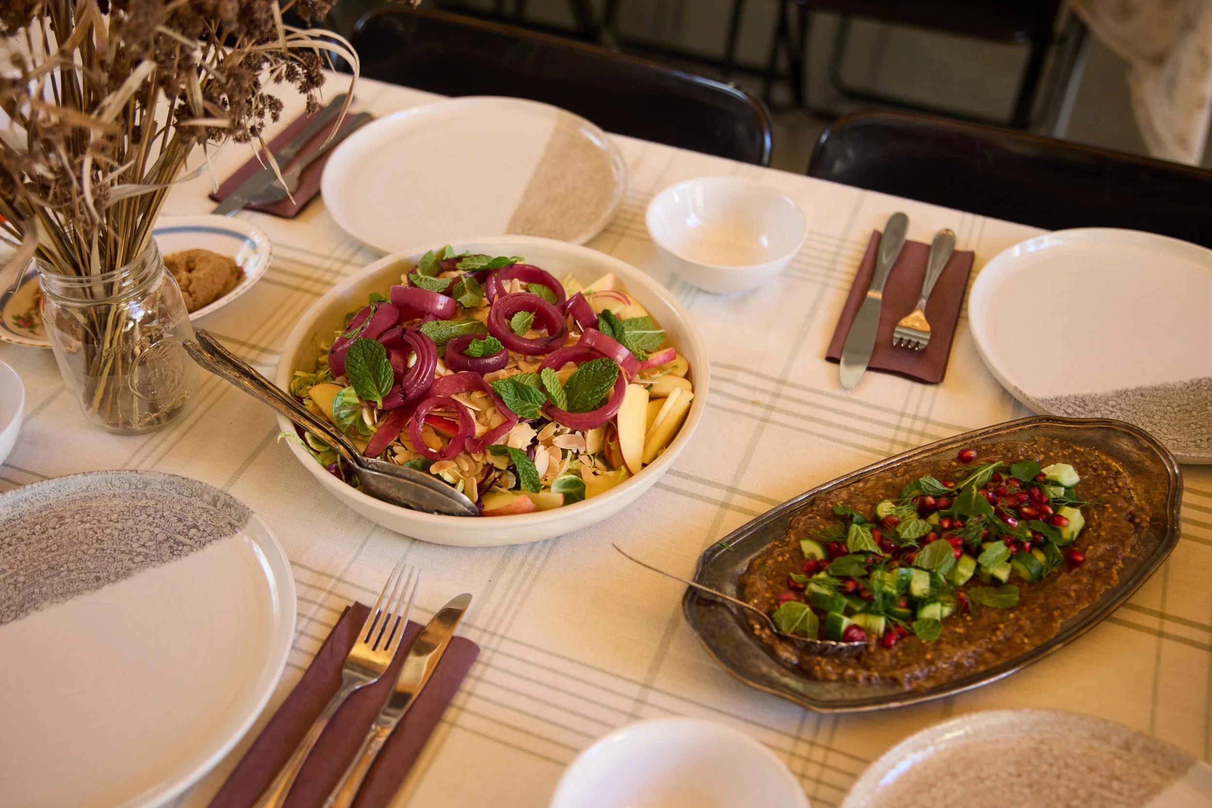 A dining table set with two plates, a salad with mint leaves and onions, a dish with cucumbers and pomegranate seeds, empty bowls, plates, silverware, and a jar of dried herbs or flowers.