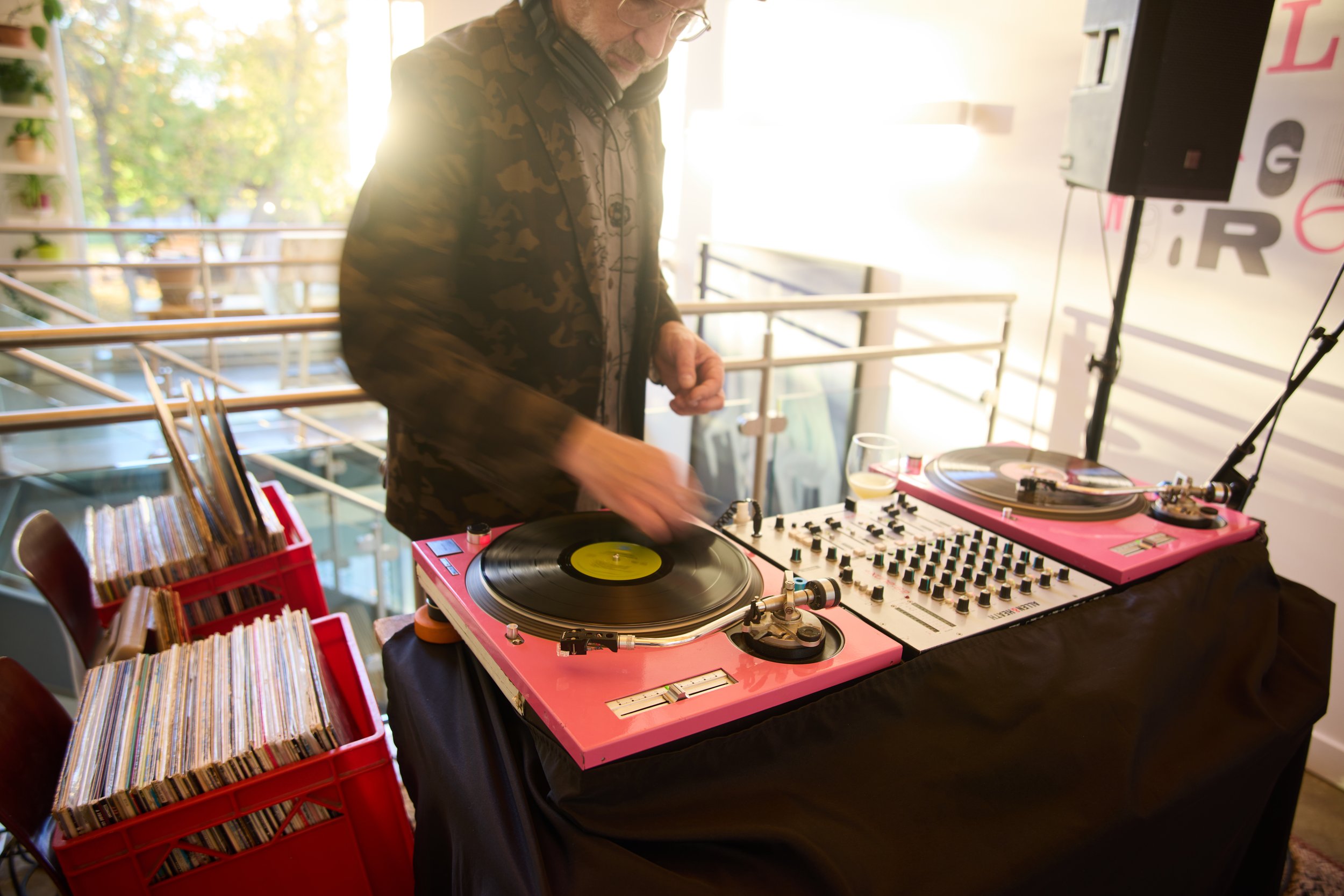 A DJ playing music on turntables and a mixer at an indoor event, with vinyl records stored in red crates nearby.