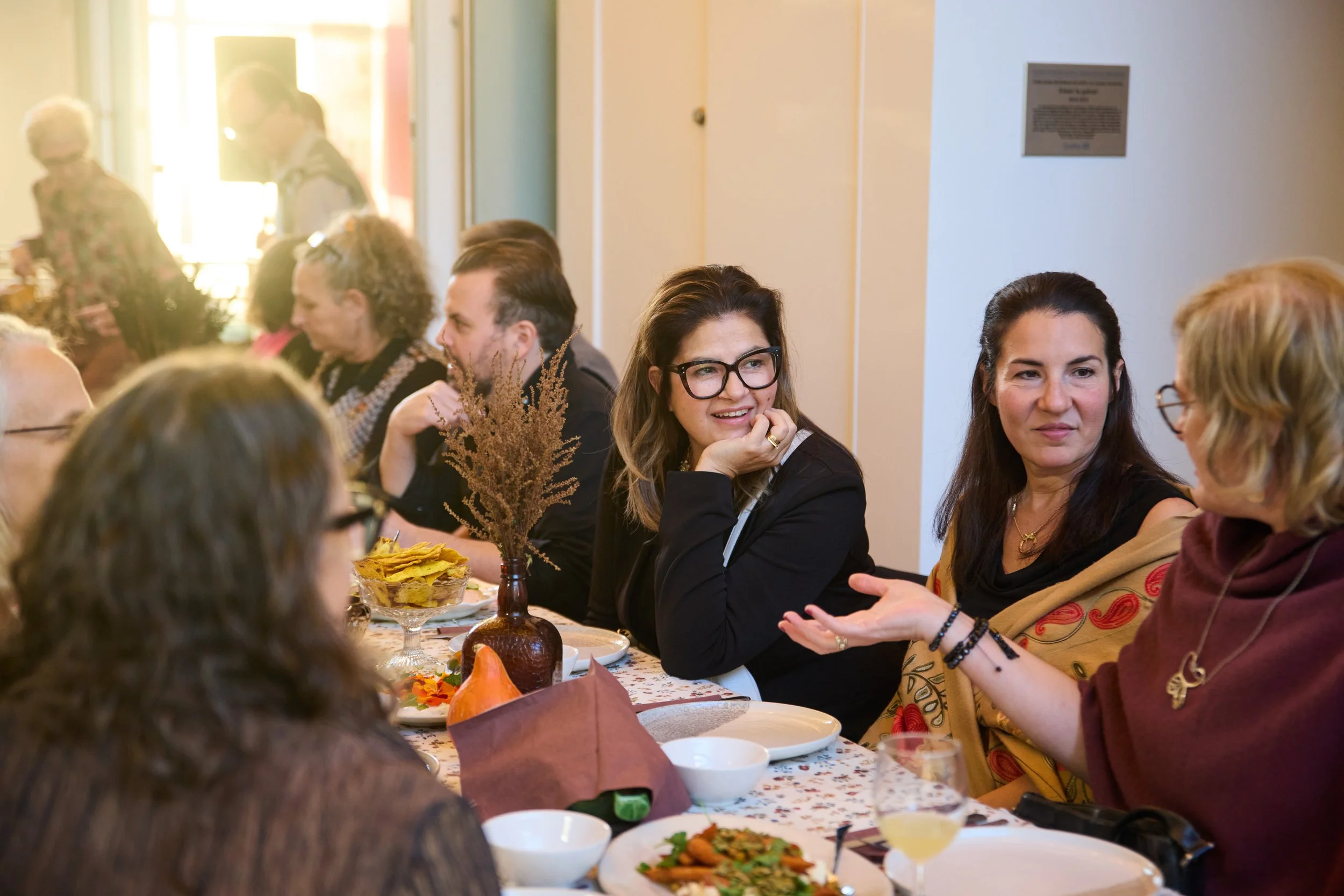 People gathered around a long table engaged in conversation at a social event or dinner, with food and drinks on the table.