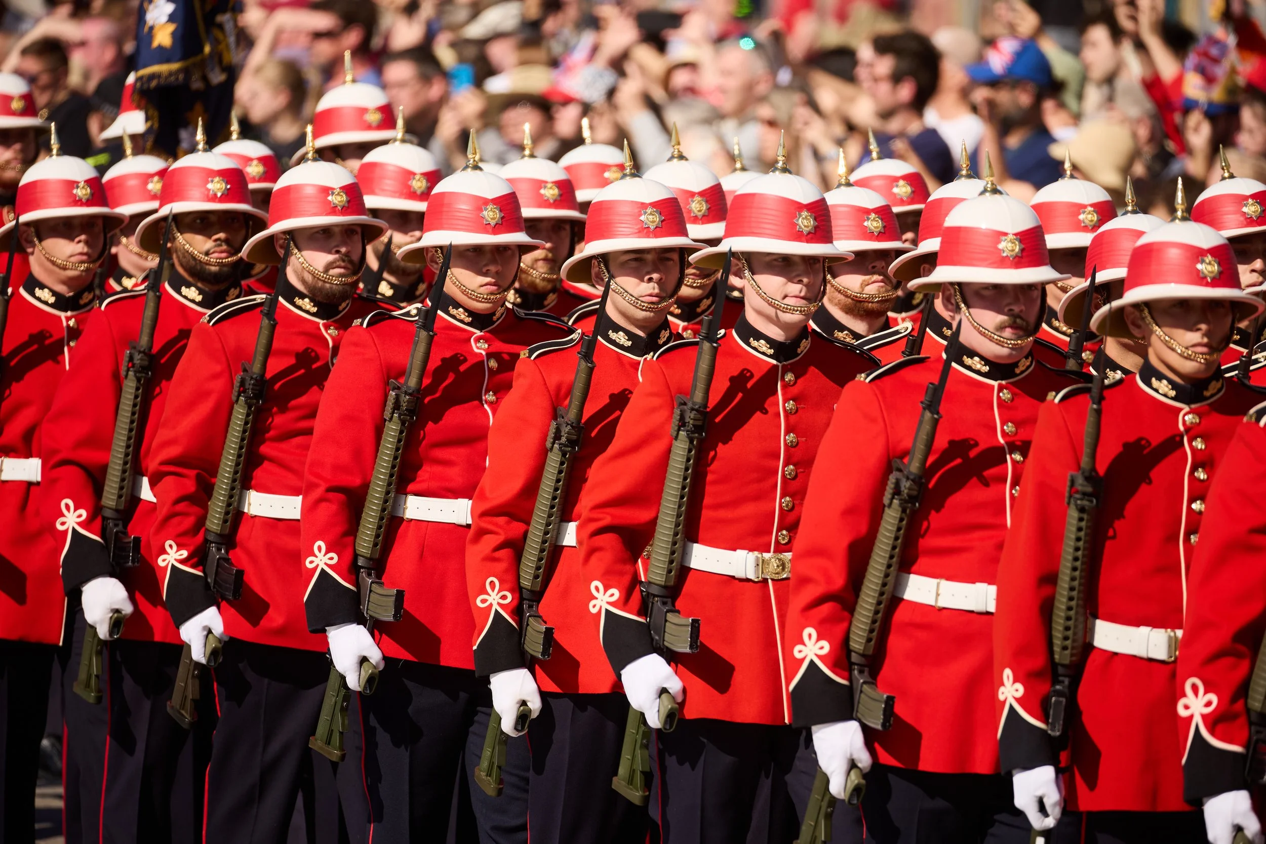 A group of ceremonial soldiers dressed in red uniforms, white helmets with red bands, and carrying rifles, participating in a parade with a crowd of onlookers in the background.