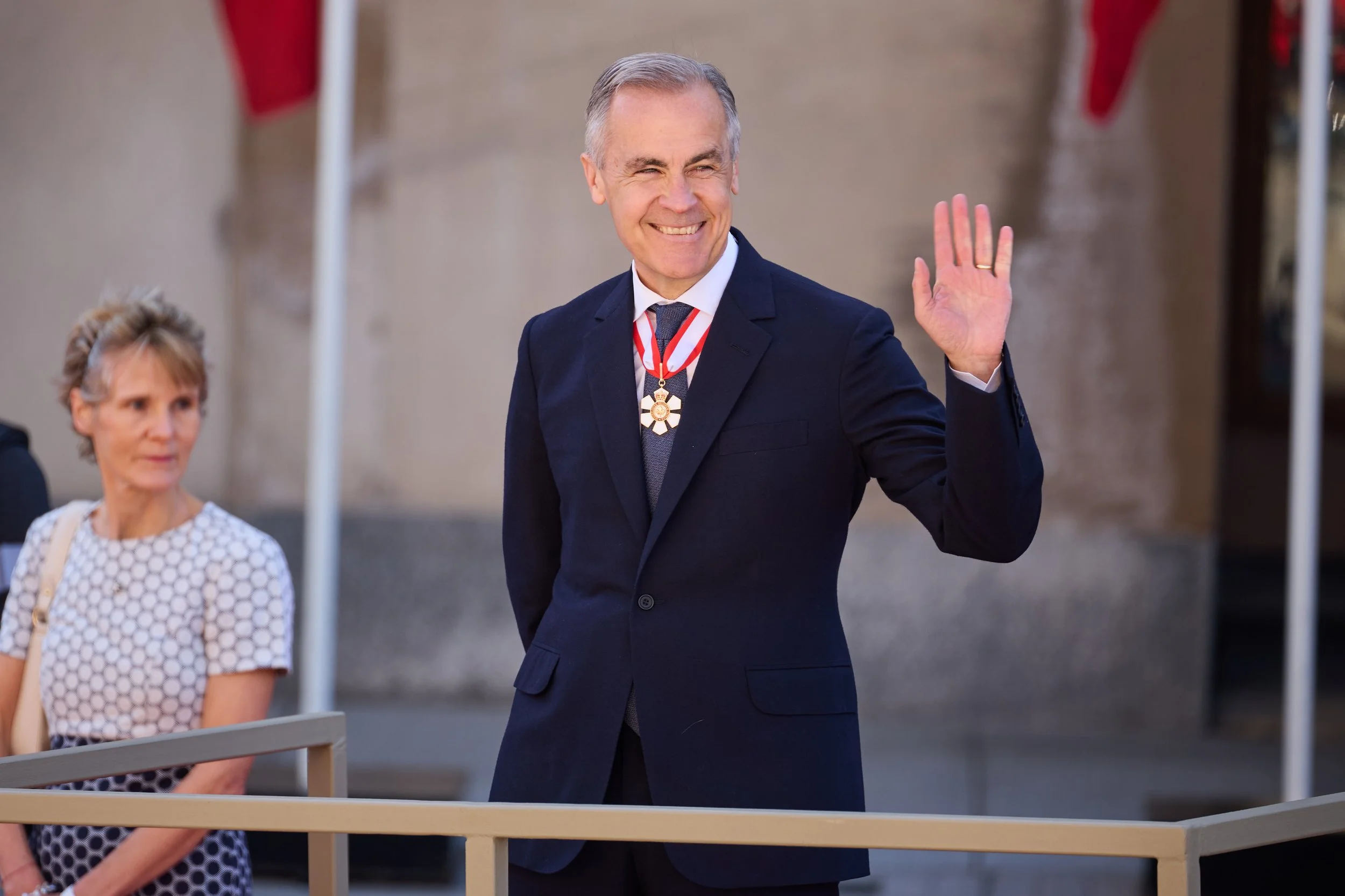 A smiling older man in a dark suit, white shirt, and tie, wearing a medal, waving with his right hand during an outdoor event. A woman with short light hair and a polka-dot dress is seated nearby watching.